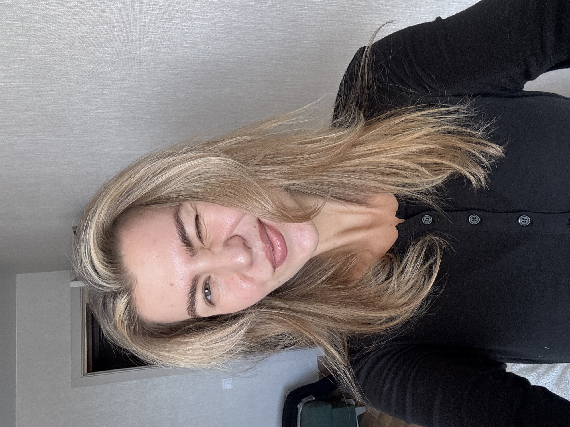A portrait-style image, rotated 90 degrees clockwise, features a young woman with long, wavy blonde hair, smiling and winking with her right eye. She is positioned as if lying down and looking up, with her head tilted slightly. Her expression is playful and friendly.
The woman has natural-looking eyebrows and her lips are glossy. Her blonde hair has lighter highlights, framing her face and cascading around her shoulders. A small red blemish is visible on her forehead. She is wearing a fitted black long-sleeved top, identifiable by several small, dark, round buttons running down the front of the garment on her chest.
The background consists primarily of a light grey wall with a subtle textured pattern. Behind her left shoulder, a rectangular frame, possibly of a window or doorway, is visible in a matching light grey tone, with a dark opening beyond it. In the bottom right corner of the full image, a glimpse of white, textured fabric (possibly a blanket) and a dark green object can be seen. There is no visible text in the image.