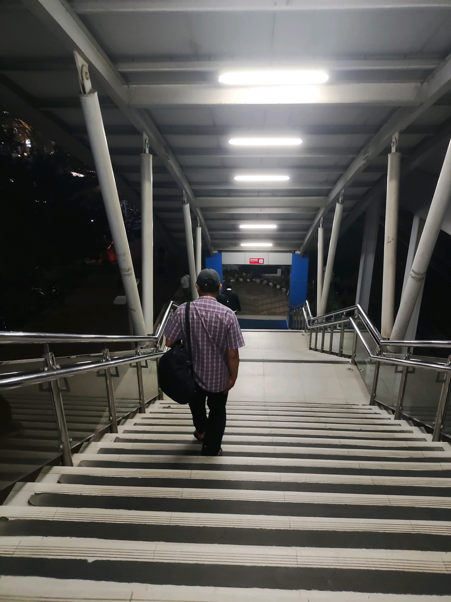 An individual wearing a plaid shirt, dark pants, and a baseball cap walks down a wide, illuminated staircase. They are carrying a dark duffel bag over their left shoulder. The staircase is covered by a metal and white-paneled walkway supported by angled white pillars. Fluorescent lights are mounted on the ceiling of the walkway. The stairs have a striped pattern of dark and light gray treads. On the right side of the stairs, a metal railing with glass panels lines the walkway. In the background, at the bottom of the stairs, there is a blue wall with a red sign that reads "CLOSED". The scene appears to be a pedestrian overpass in East Jakarta, Indonesia, likely near a transportation hub or public facility, given the "CLOSED" sign and the architectural style. The surrounding environment visible on the left of the walkway is dark, suggesting nighttime or a dimly lit area with some foliage.