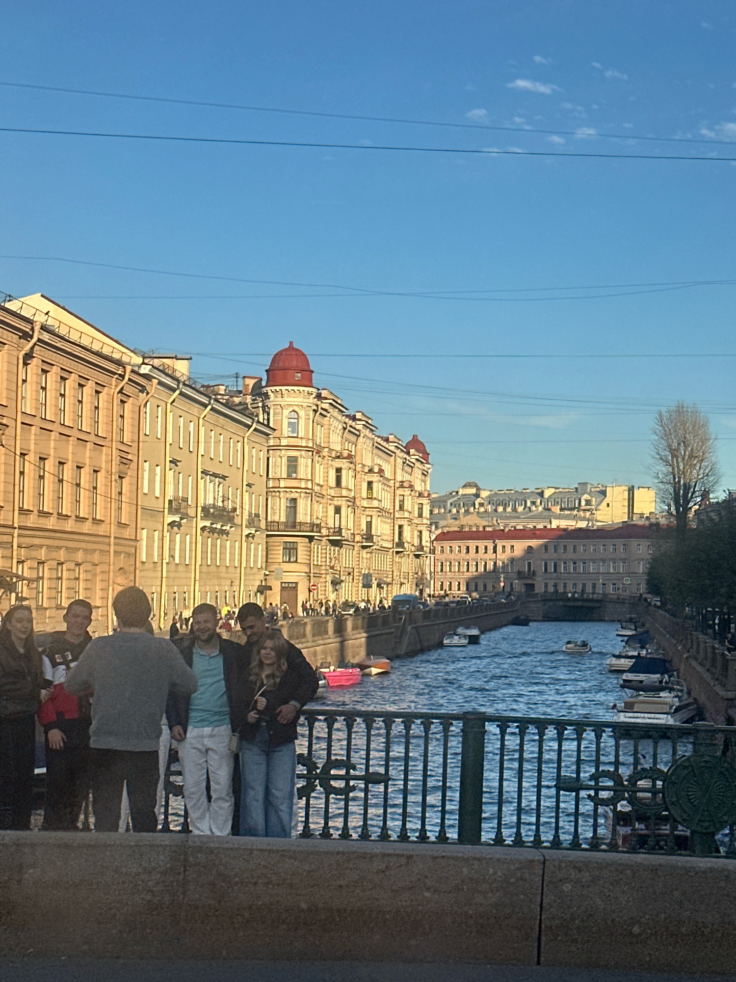The image depicts a group of people standing on a bridge overlooking a canal in Saint Petersburg, Russia. The canal is lined with ornate buildings, characteristic of the city's architecture, with a prominent red dome visible on one of the buildings. Several boats are navigating the canal, and the sky above is a clear blue with a few wisps of clouds. The people on the bridge appear to be casually dressed, with some engaging in conversation and others looking out at the scenery. The overall scene suggests a pleasant day in a historic urban setting.