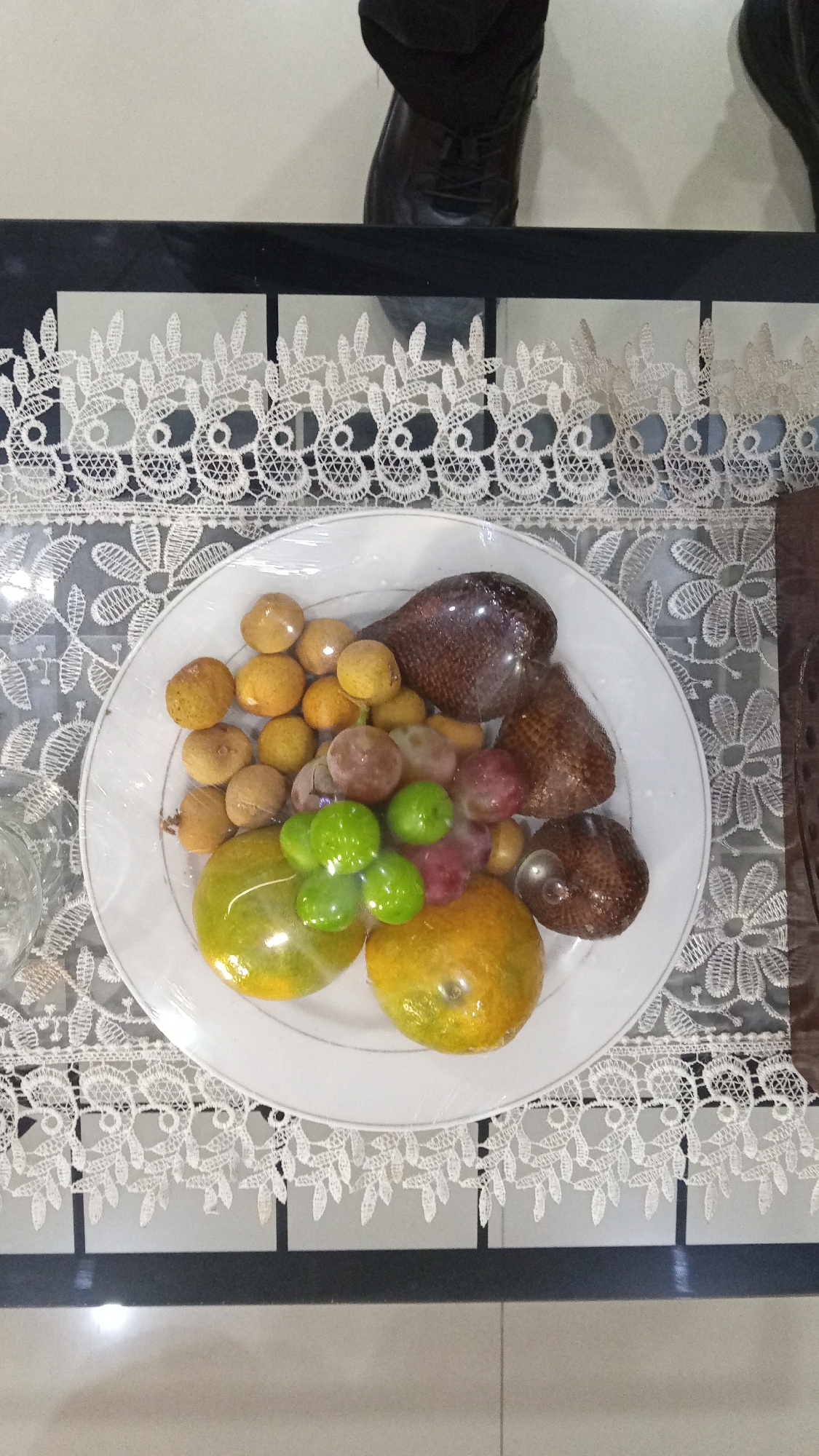 A white plate filled with assorted fruits, including salak, longan, grapes, and oranges, is placed on a table covered with a white lace tablecloth. The plate and fruits are wrapped in clear plastic. Two dark leather shoes are visible at the top of the image, suggesting someone is standing near the table. The background is a neutral color. The image is likely taken indoors in a home or event setting.