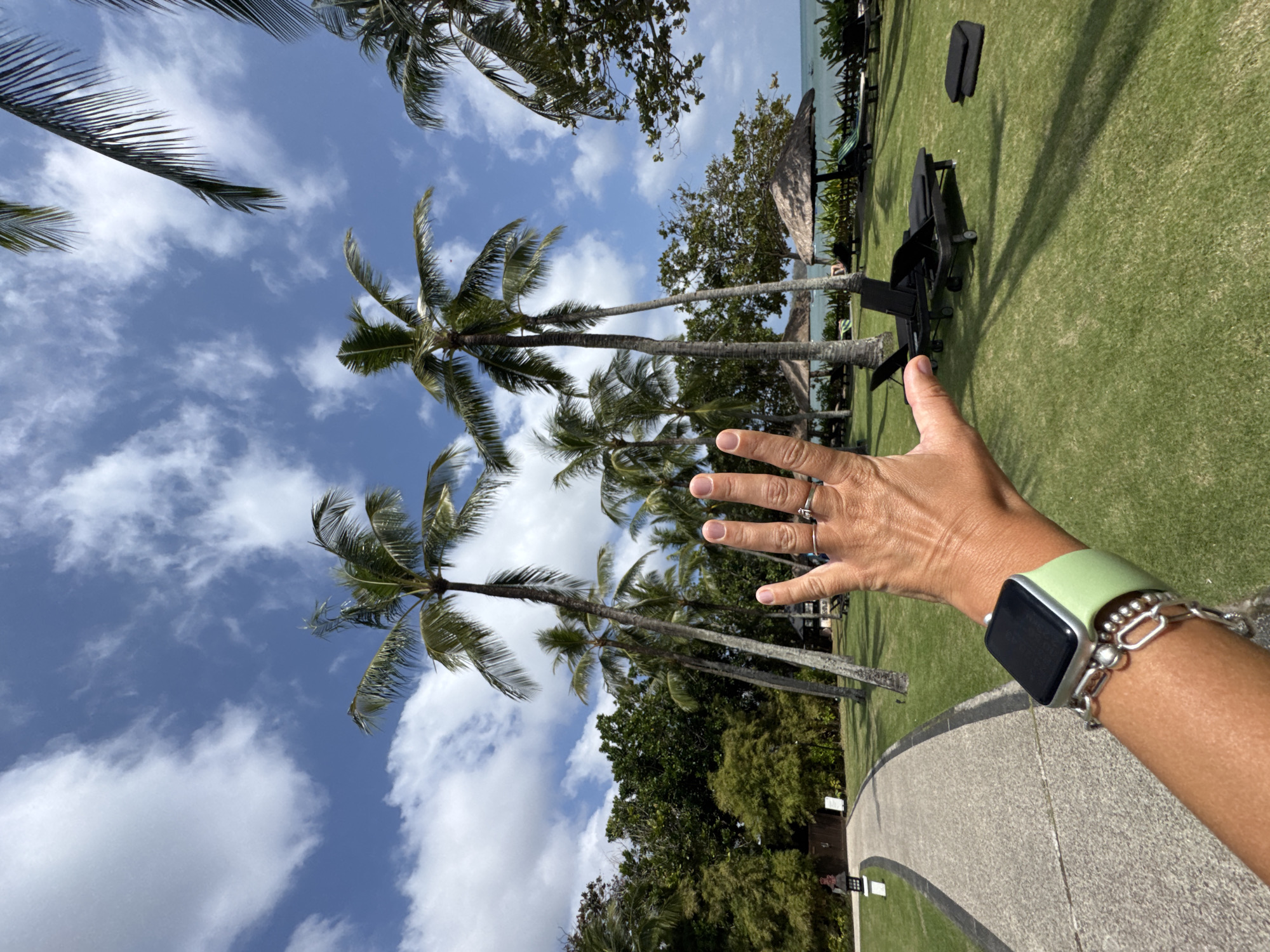 The image shows a person's hand in the foreground, with fingers extended. The hand has a silver ring on one finger and a watch on the wrist. The background depicts a tropical scene with palm trees, lush green grass, and lounge chairs. A pathway curves through the grass. In the distance, there is a body of water, likely the ocean, and umbrellas providing shade over lounge chairs. The sky is blue with scattered white clouds. The overall impression is a resort or beach setting. Based on the context of Kuta Selatan, Indonesia, this scene likely depicts a resort area with typical tropical amenities.