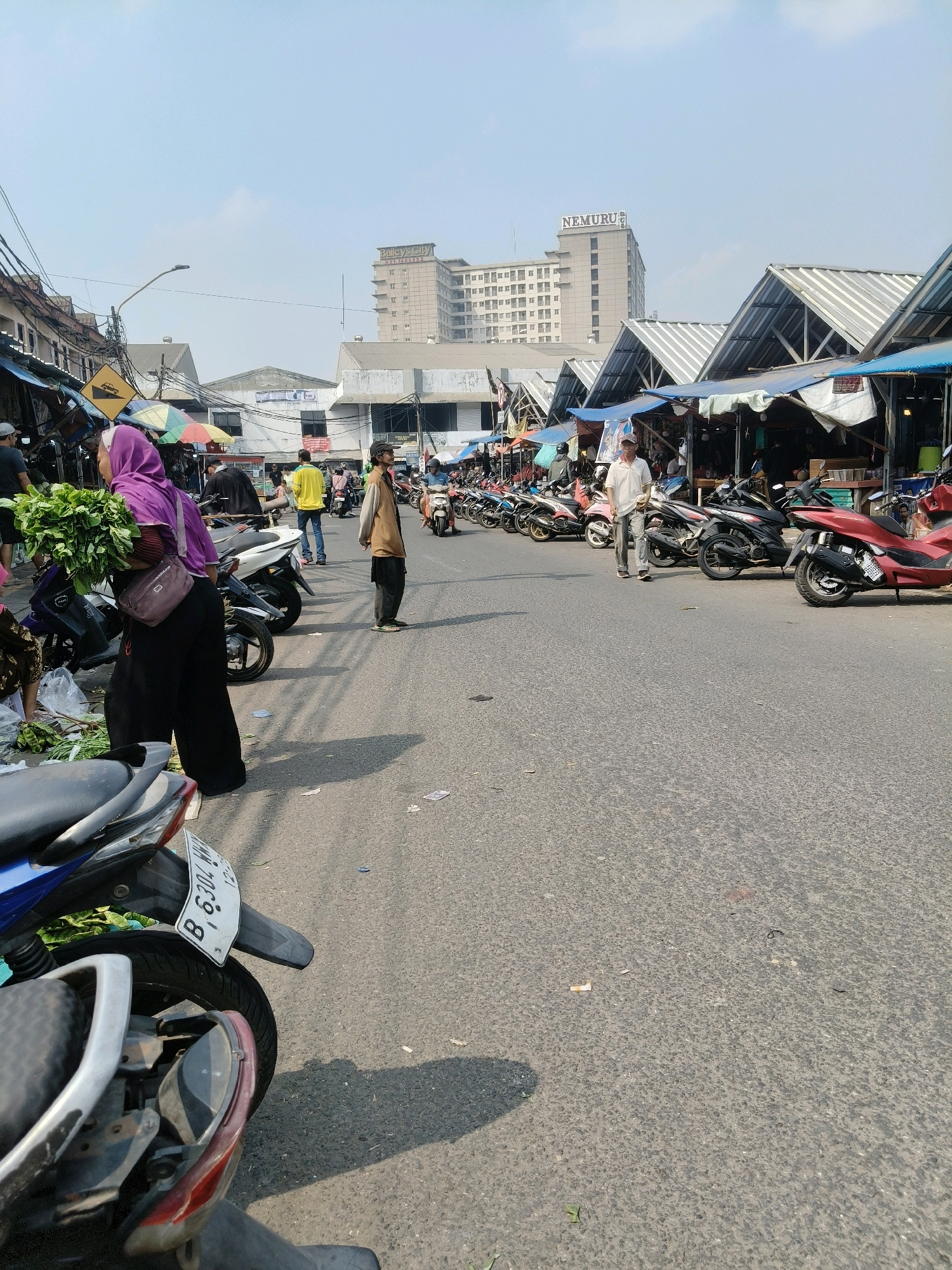 The image captures an outdoor market scene in South Tangerang, Indonesia, on a clear day. A paved road runs through the center, lined with stalls covered by various awnings and tarps, suggesting a bustling marketplace. Various motorcycles are parked along the side of the road, indicating a mode of transportation for vendors and shoppers.
In the foreground, a woman wearing a purple hijab and black clothing carries a large bunch of leafy green vegetables. To her right, a man in a tan jacket and dark pants stands facing forward. Further down the road, another man in a white shirt and light-colored pants walks away from the camera, looking towards the stalls. A few other individuals are visible in the background, including a person in a yellow shirt walking down the street.
The environment appears somewhat urban, with buildings visible in the distance. One prominent building displays a sign that reads "NEMURU," and another building, taller and lighter in color, shows signs with text that is difficult to decipher. The sky is bright and blue, with a few wisps of clouds. The road surface shows signs of wear and tear, with some debris scattered on it. The overall impression is one of a lively, everyday market day.