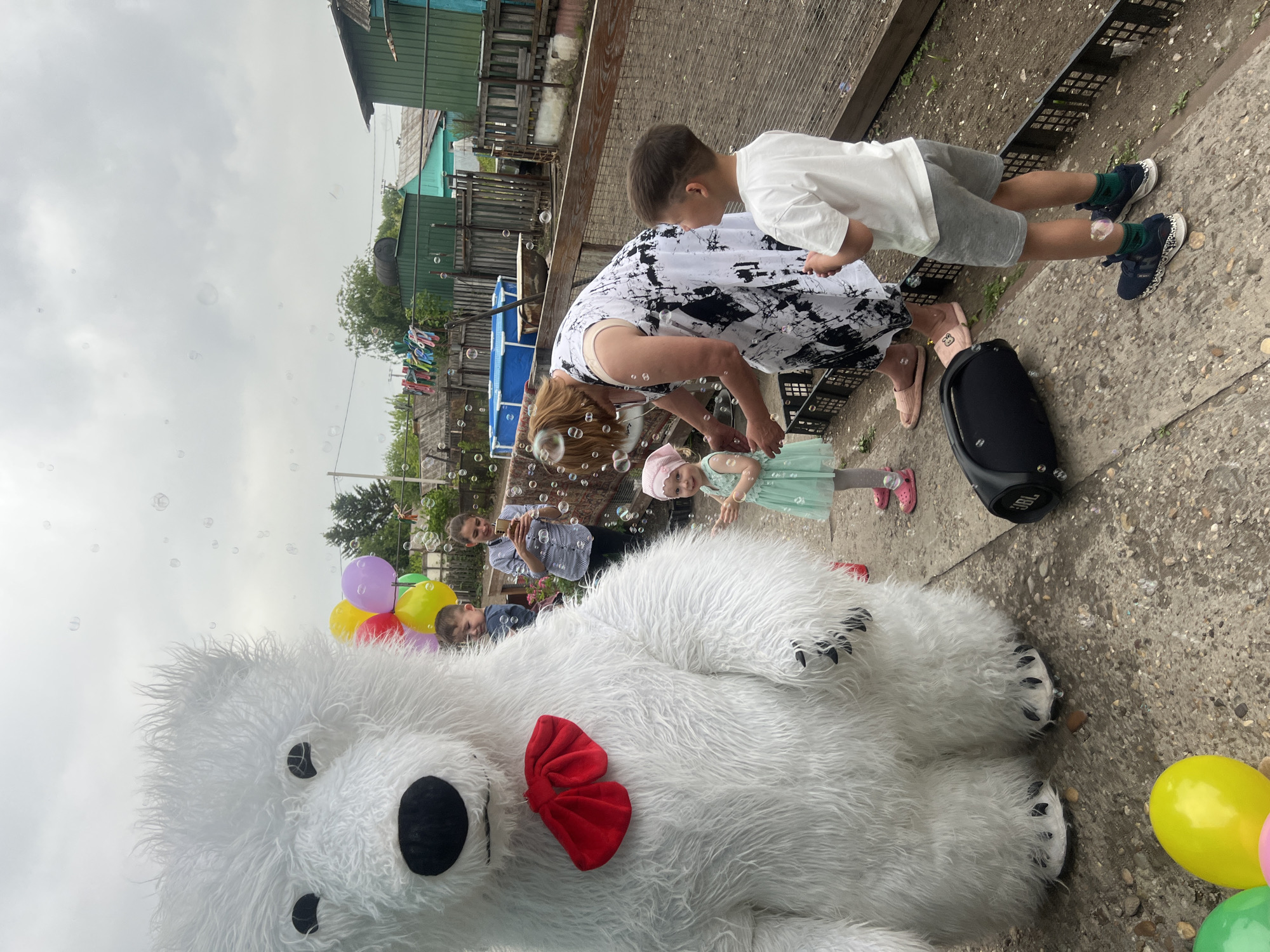 The image shows a group of people, including children, gathered outdoors in an urban location, likely in the city of Unknown, Unknown. A large white polar bear costume, complete with a red bow tie, dominates the foreground. Scattered around are colorful balloons and numerous soap bubbles floating in the air, suggesting a celebratory atmosphere. A woman in a black and white dress is bending down, possibly interacting with a young girl in a light green dress and pink hat. A boy is standing nearby, observing. In the background, several adults are standing, with one appearing to be taking a photo or video with a mobile phone. The background also reveals residential buildings, adding to the urban setting. A black JBL speaker sits on the ground near the woman and children. The ground appears to be concrete or asphalt with some pebbles. The overall scene is lively and celebratory.