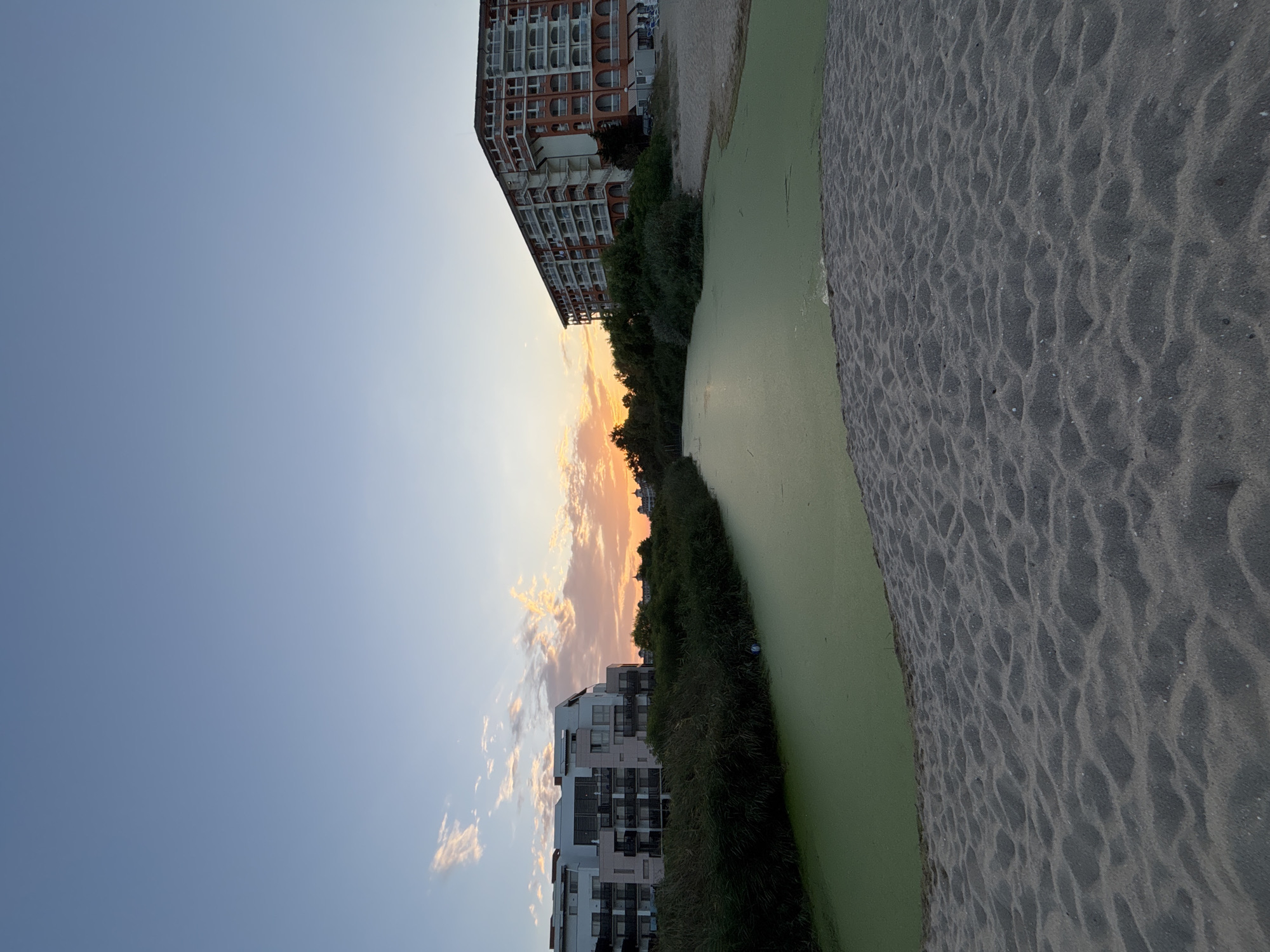 The image shows a body of water covered in green algae, with sandy ground in the foreground. In the background, there are two buildings, likely hotels, and a line of trees. The sky is clear with a few clouds tinged with the orange and pink hues of a sunset. The scene is quiet and peaceful, suggesting it might be a tourist resort area in Obzor, Bulgaria, during the evening.