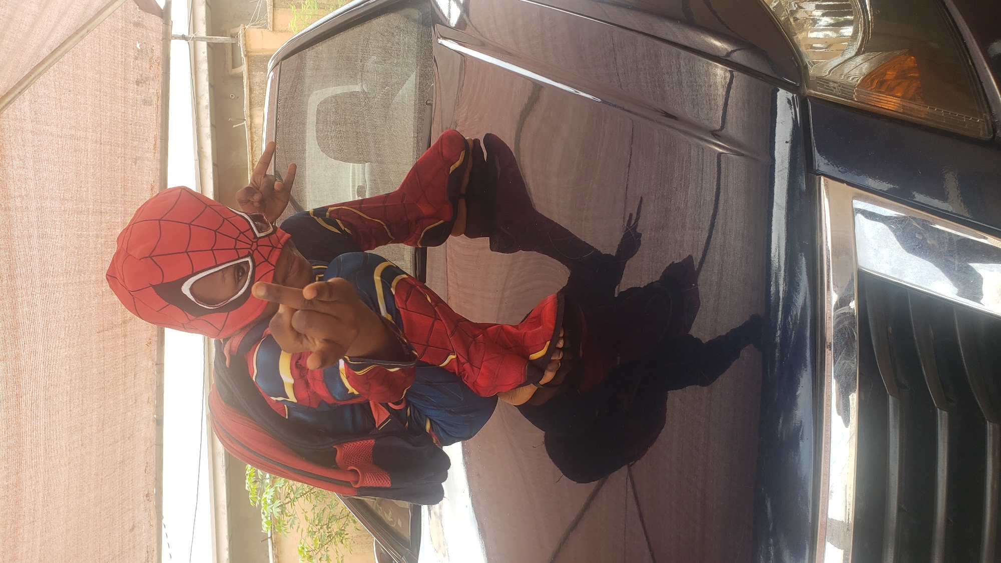 A person dressed as Spider-Man poses against the shiny surface of a dark-colored vehicle, likely a car, in an outdoor setting. The individual is wearing a full Spider-Man costume with a red and blue pattern, a mask, and footwear resembling boots. They are making gestures with their hands; one hand is displaying a 'rock on' sign while the other is extending the middle finger. The reflection of the person can be seen on the car's surface. The location is identified as Sabon Tasha, Nigeria, based on the geotag information associated with the image. The environment appears to be a semi-enclosed space, possibly a garage or sheltered area, with a roof and partial wall visible.