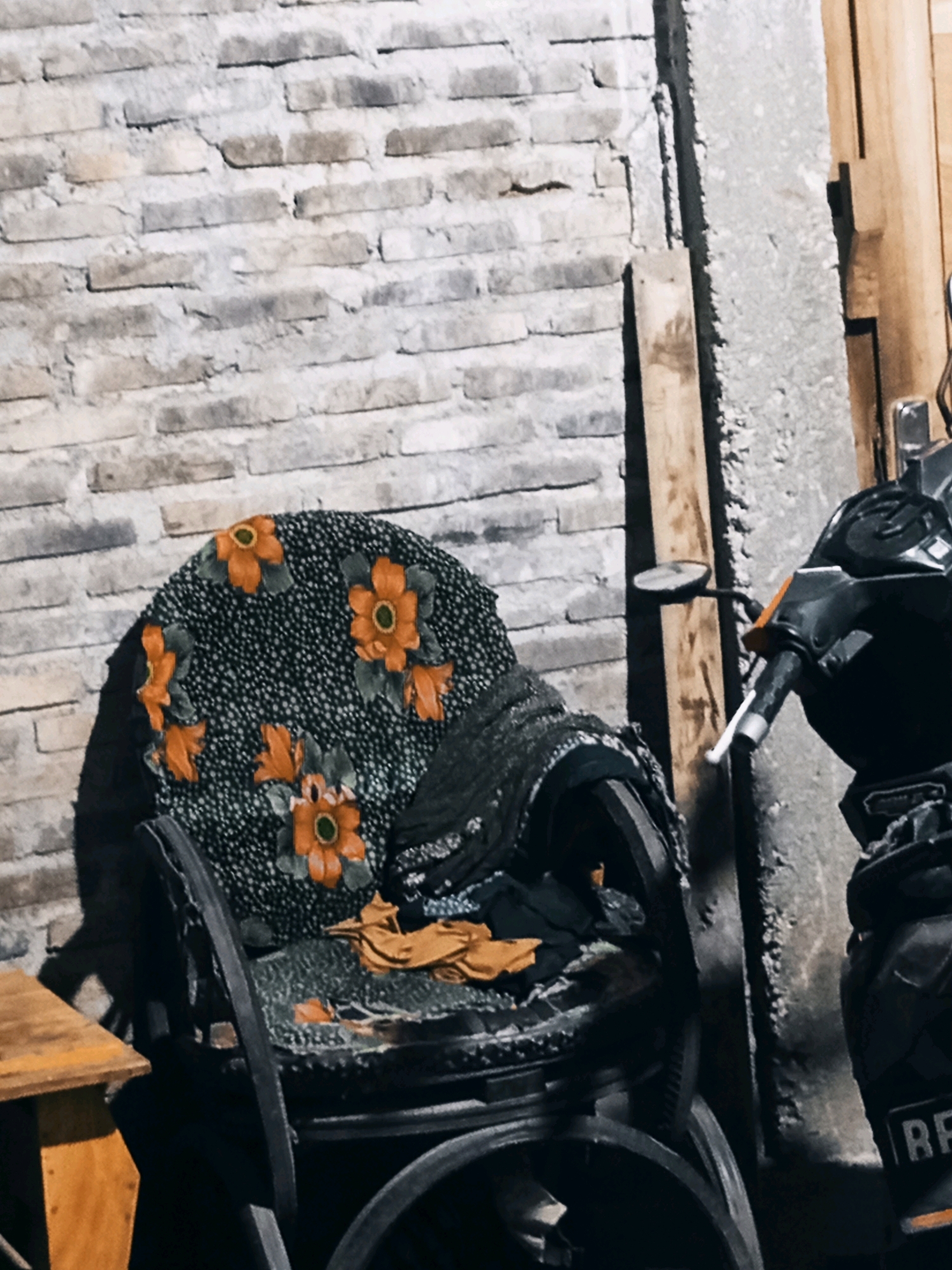 The image depicts a scene in Karang Sari, Indonesia. In the foreground, a weathered armchair with a floral patterned fabric is visible, showing signs of wear and tear. To the right of the armchair, a motorcycle is parked. The background consists of a textured brick wall. The lighting suggests it is either dusk or night.