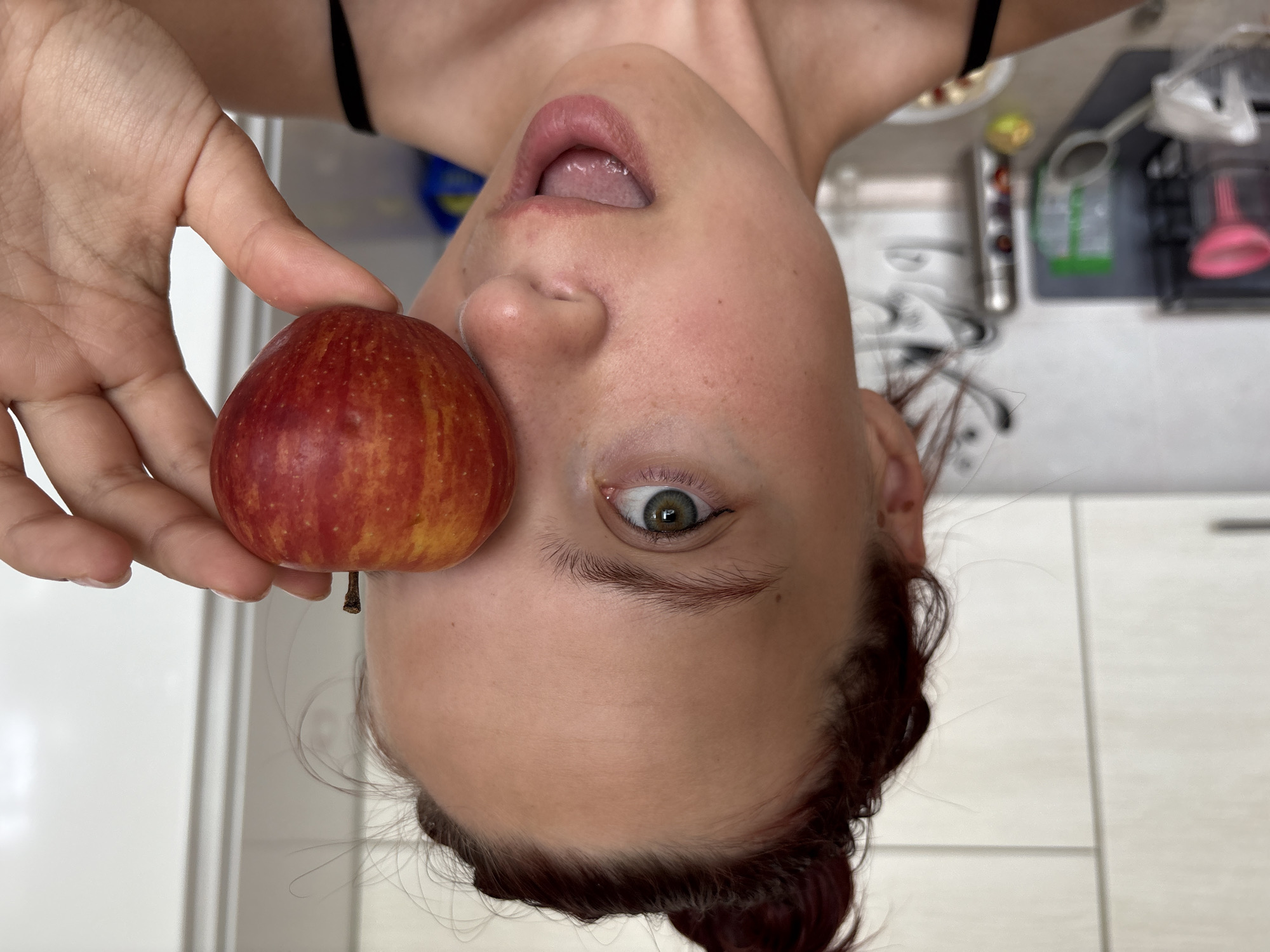 An upside-down view features a woman holding a red apple in front of her face. Her mouth is open, and one eye is visible. The background shows a kitchen counter with various items. The location is mentioned as Прикамский, Russia, but there is no confirmation from the visual data to substantiate this fact.
