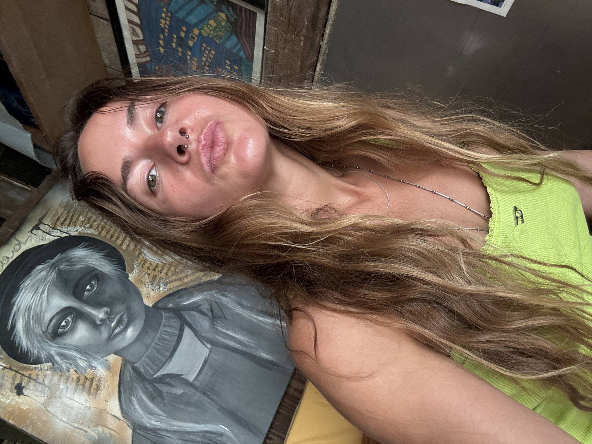 A woman with long, wavy blonde hair is taking a selfie in what appears to be a room with artwork on the walls. She is wearing a bright green tank top with a silver necklace. She has a septum piercing and is making a pouty face towards the camera. In the background, a painting of a person wearing a hat and a sweater is visible, as well as another abstract painting with blue and yellow geometric shapes. The room seems rustic, with exposed wooden beams and shelves.