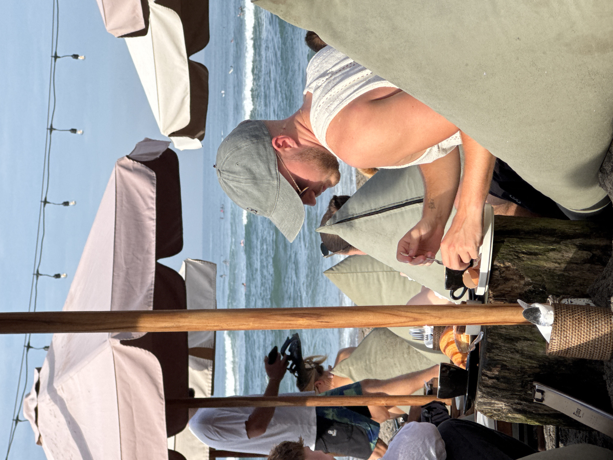The image shows an outdoor scene in North Kuta, Indonesia, likely a restaurant or beachside café. A fair-skinned man with a beard, wearing a grey baseball cap and white tank top, is seated at a table. He is interacting with items on the table, which include a white plate and a dark-colored cup. Other people are at the table, including one person taking a photo and another with fair-colored hair. Umbrellas provide shade. The ocean is visible in the background.