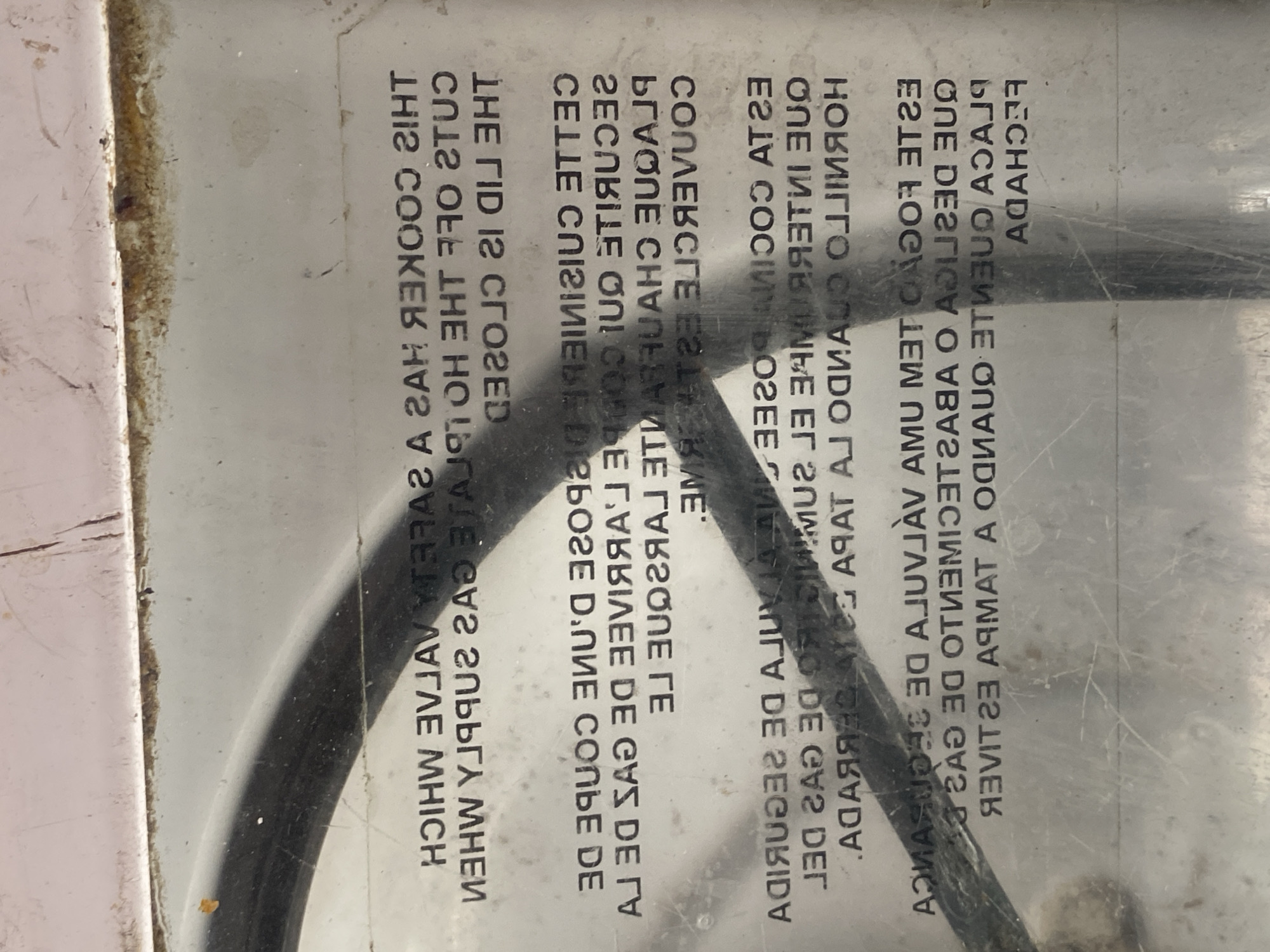 This is a close-up, reflective photograph of a metal appliance, likely a stovetop or oven, with text printed on its surface. The text is in multiple languages, appearing reversed due to the reflective nature of the surface. There are no people, animals, or recognizable city landmarks visible. The background is out of focus and appears to be an indoor setting. The text is the primary subject of the image, and it contains instructions or warnings related to the appliance's operation. The photograph's reflective quality creates distorted shadows and highlights, obscuring some of the details. The location of Jauro Yinu, Nigeria, is not determinable from the image content.