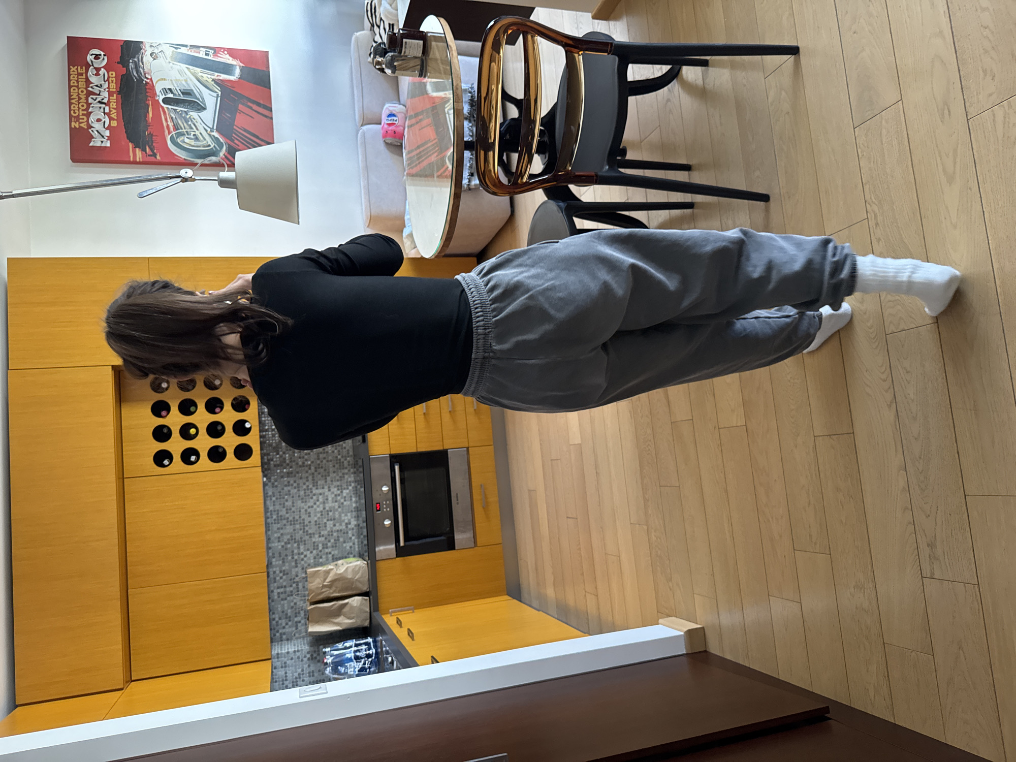 An indoor scene captured in an apartment, likely located in Warsaw, Poland, showcasing a modern open-plan kitchen and living area. A person with long, dark brown hair, wearing a black long-sleeved top, grey sweatpants, and white socks, stands with their back to the camera. They are looking down towards the kitchen counter, seemingly engaged in an activity or task.
The kitchen area features distinctive yellow wooden cabinetry, a grey tiled backsplash, and a built-in Bosch oven with a digital display. An integrated wine rack, with several empty slots, is visible within the upper cabinets. On the counter, two brown paper bags and a container with a blue lid are present.
Adjacent to the kitchen is a living or dining space, furnished with a light-colored sofa, a round glass-top table, and a contemporary chair with a glossy amber-colored backrest and black legs. A floor lamp with an adjustable arm and a light-colored shade is positioned near the sofa. On the table, a Pepsi can is visible.
A vintage-style poster depicting a race car hangs on the wall, with text that reads "2E GRAND PRIX AUTOMOBILE", "MONACO", and "8 AVRIL 1930". The flooring throughout the space consists of light-colored wooden planks. The overall aesthetic is modern and minimalist, with natural light subtly illuminating the interior.