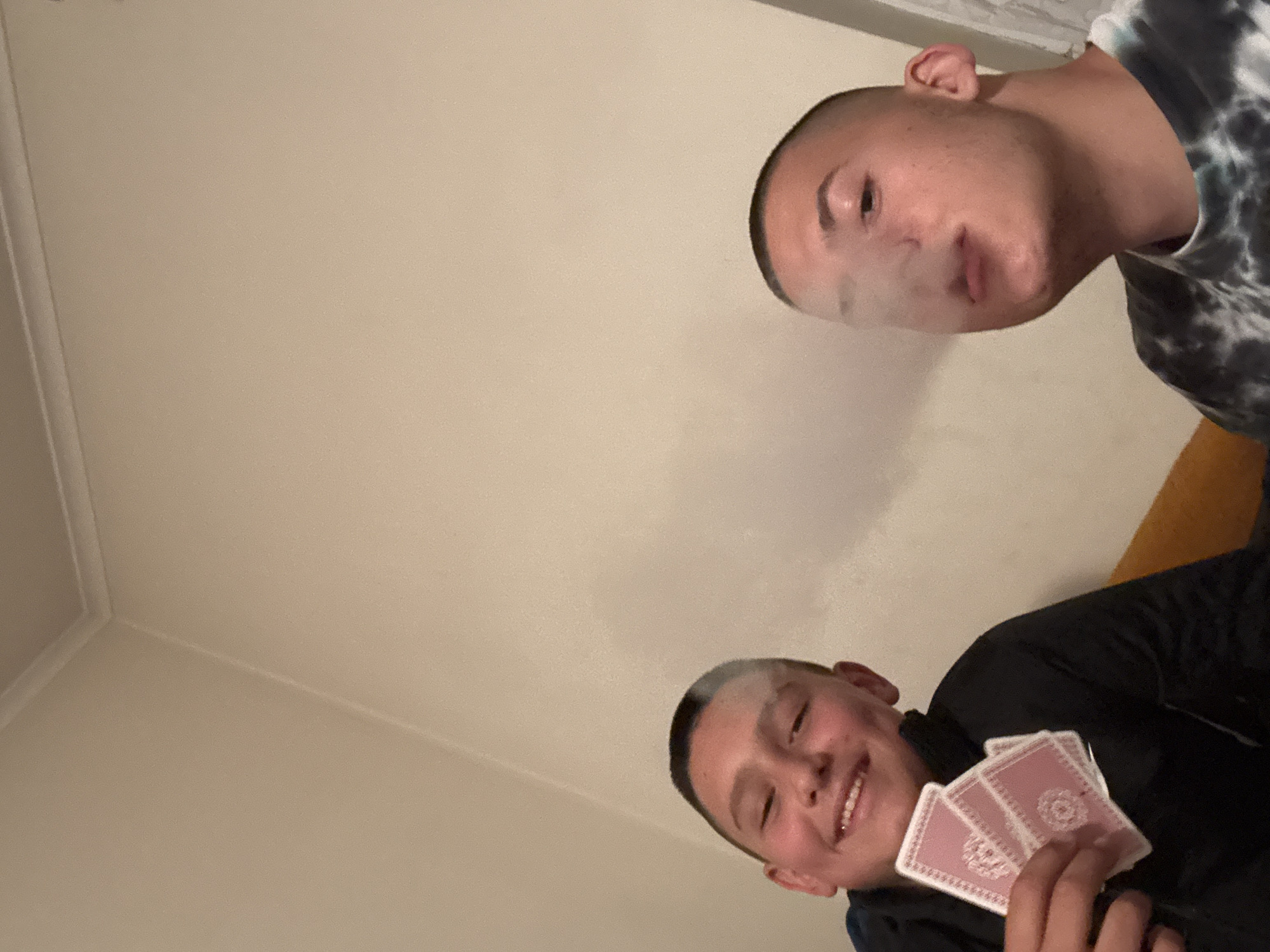 The image depicts two young men in an interior setting, likely a room. One man is visible in the upper right, exhaling vapor. The other man is holding a hand of playing cards and smiling. The background shows a beige wall. The location in the city of Goritsa, Bulgaria, is not supported by the visual information in the image.