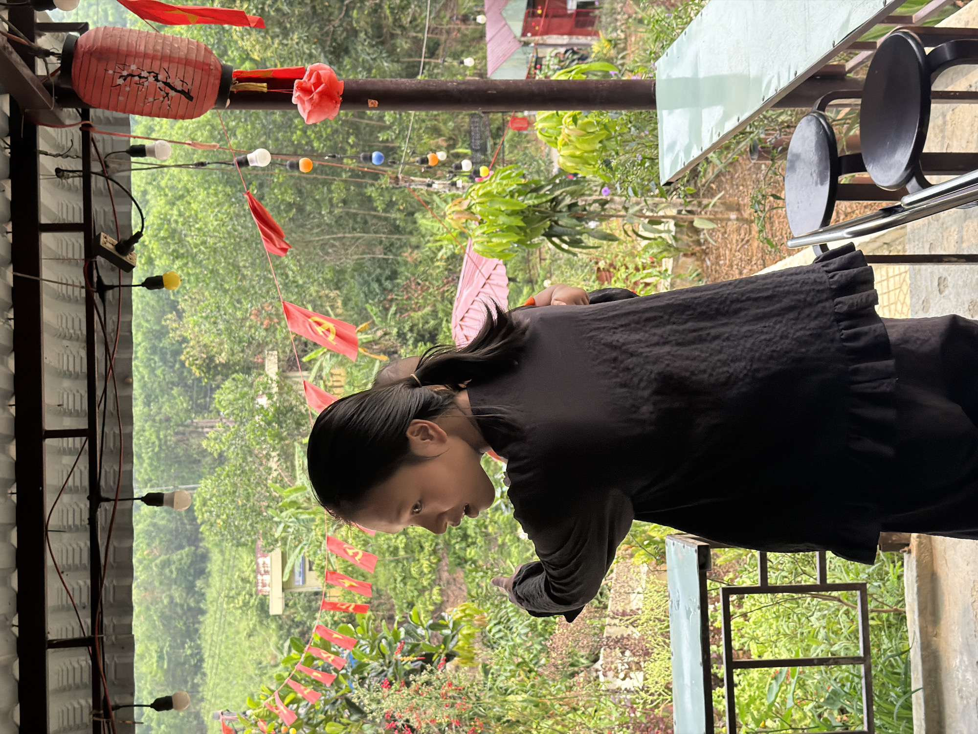A medium shot shows a Vietnamese woman in a black shirt pointing down with her right hand. The camera angle is such that the viewer sees the woman's left profile. The scene is primarily outdoors, as indicated by the vegetation and lighting. A decorative red lantern with black floral accents hangs above the woman, along with a string of lights and small red flags that are likely national flags of Vietnam. The scene gives the impression of a casual outdoor cafe or restaurant setting, with metal table and chair frames. The background is rich with greenery, indicating a natural or garden setting, and hints of structures can be seen amidst the trees. While the image does not confirm the location in Đà Nẵng, Vietnam, the presence of the Vietnamese flags and the general style of the scene suggest it could be a plausible location.