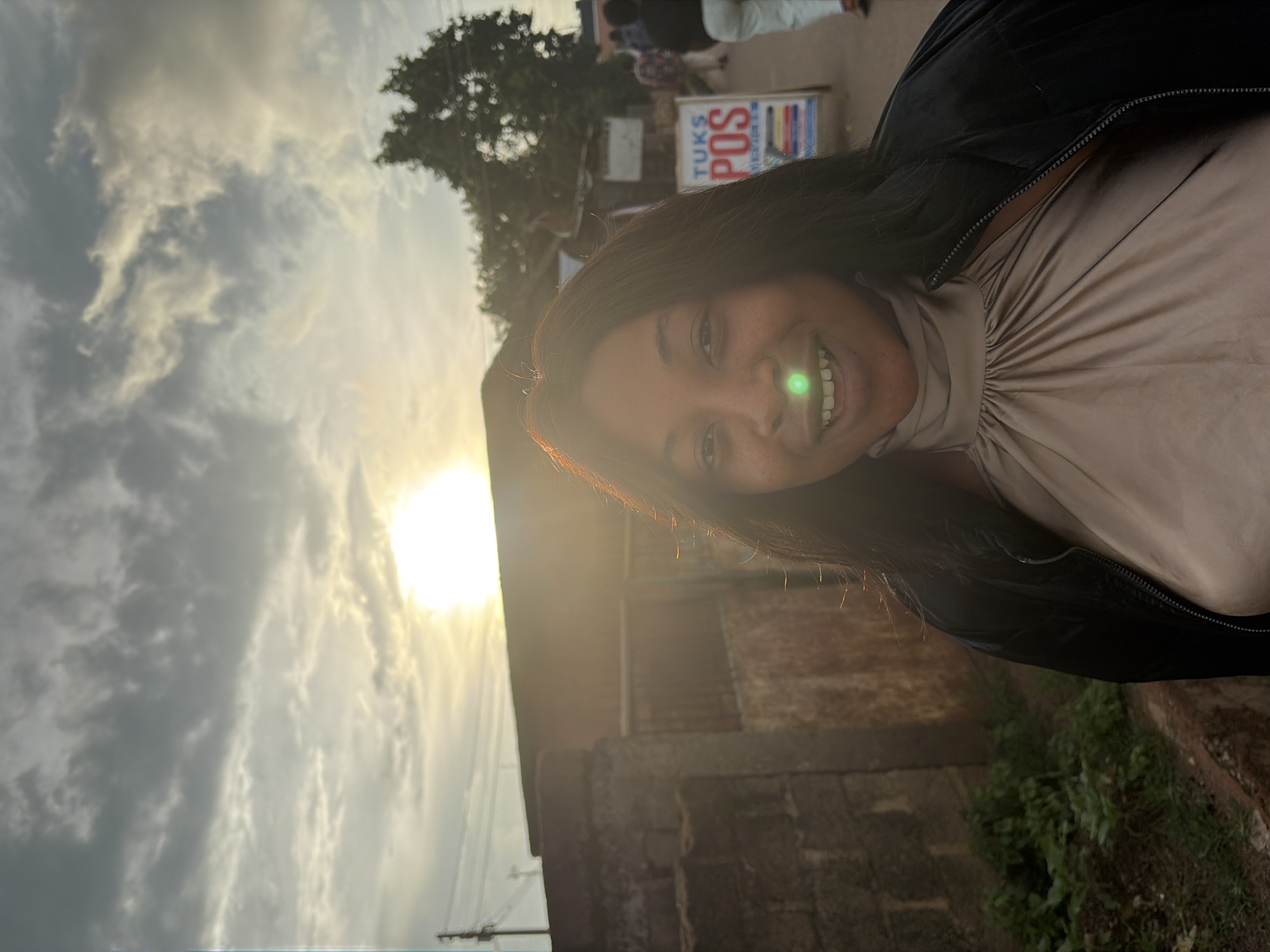 A young Black woman with dark hair smiles at the camera, her face illuminated by the setting sun. She wears a beige silk top with a high neckline and a black jacket. A green light reflects on her nose. She stands on a dirt path next to a brick wall and some greenery. In the background, a building with a corrugated metal roof is visible, along with a street with people walking and a sign that reads "TUKS POS." The sky is cloudy with the sun breaking through. The scene appears to be in an outdoor marketplace or residential area in Jos, Nigeria, given the architectural style and the signage.