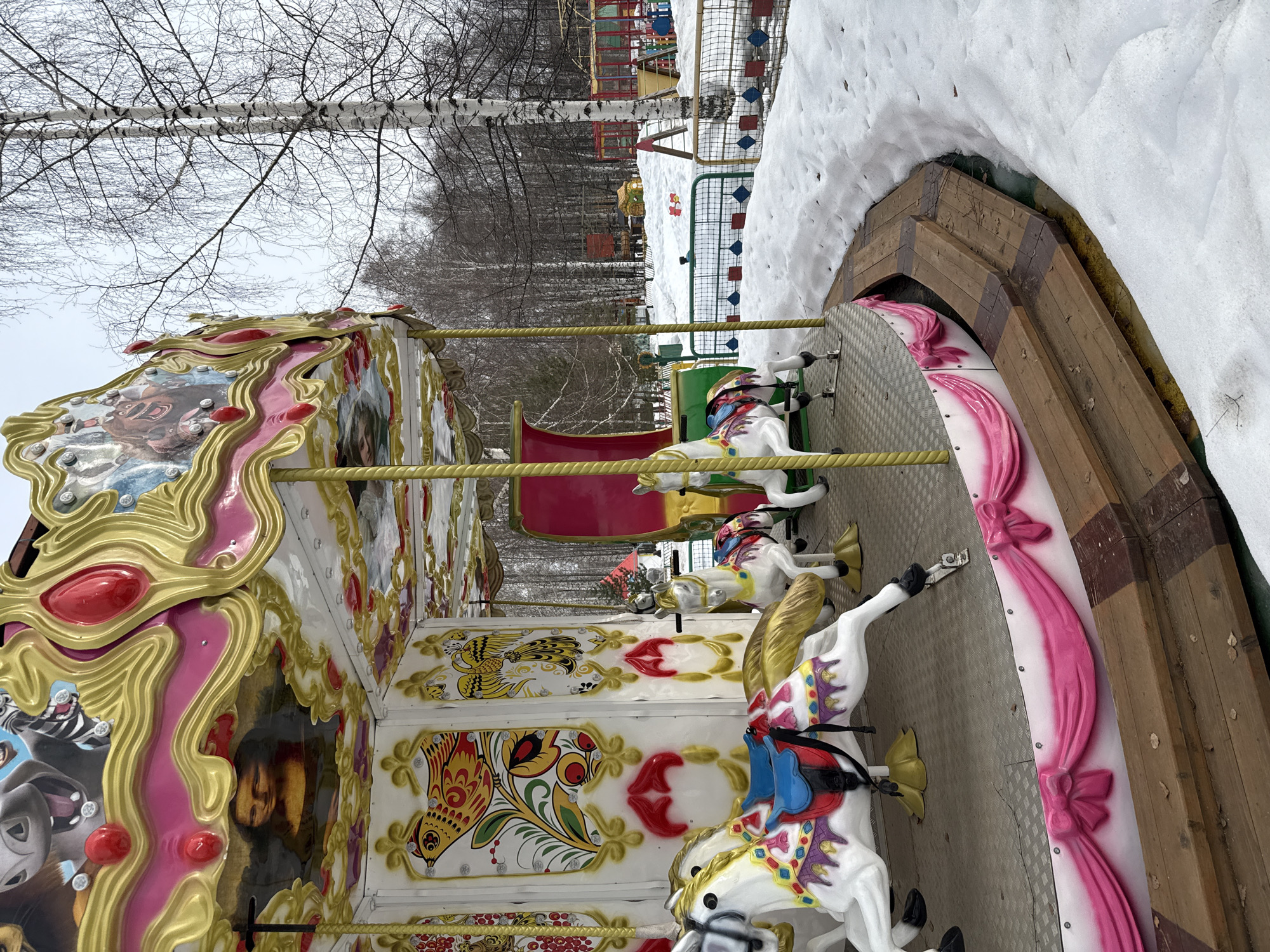 The image displays a brightly decorated carousel situated in a snowy park or amusement area within the city of Chaikovsky, Russia. The carousel, largely in the foreground, features a prominent gold and pink canopy adorned with various images, including what appears to be a rendition of the Mona Lisa and cartoon animal characters like a hippo and a zebra. The interior panels are decorated with colorful traditional Russian folk art birds and floral motifs on a white background. Several white carousel horses with multi-colored saddles and bridles, along with a single red carriage, are visible on the metal riding platform. The platform is bordered by a pink, ribbon-draped facade and a dark wooden two-step perimeter.
The ground around the carousel is covered in patches of snow, with some melted areas revealing wet earth and sparse dead leaves. To the right and in the background, a cluster of bare deciduous trees, primarily birches with their distinctive white bark, stands against an overcast sky. Beyond the trees and behind a low metal fence, additional colorful playground equipment, including slides, swings, and climbing structures, is partially visible and also covered in snow, indicating a winter or early spring setting. No individuals are present in the scene, and the carousel appears stationary.