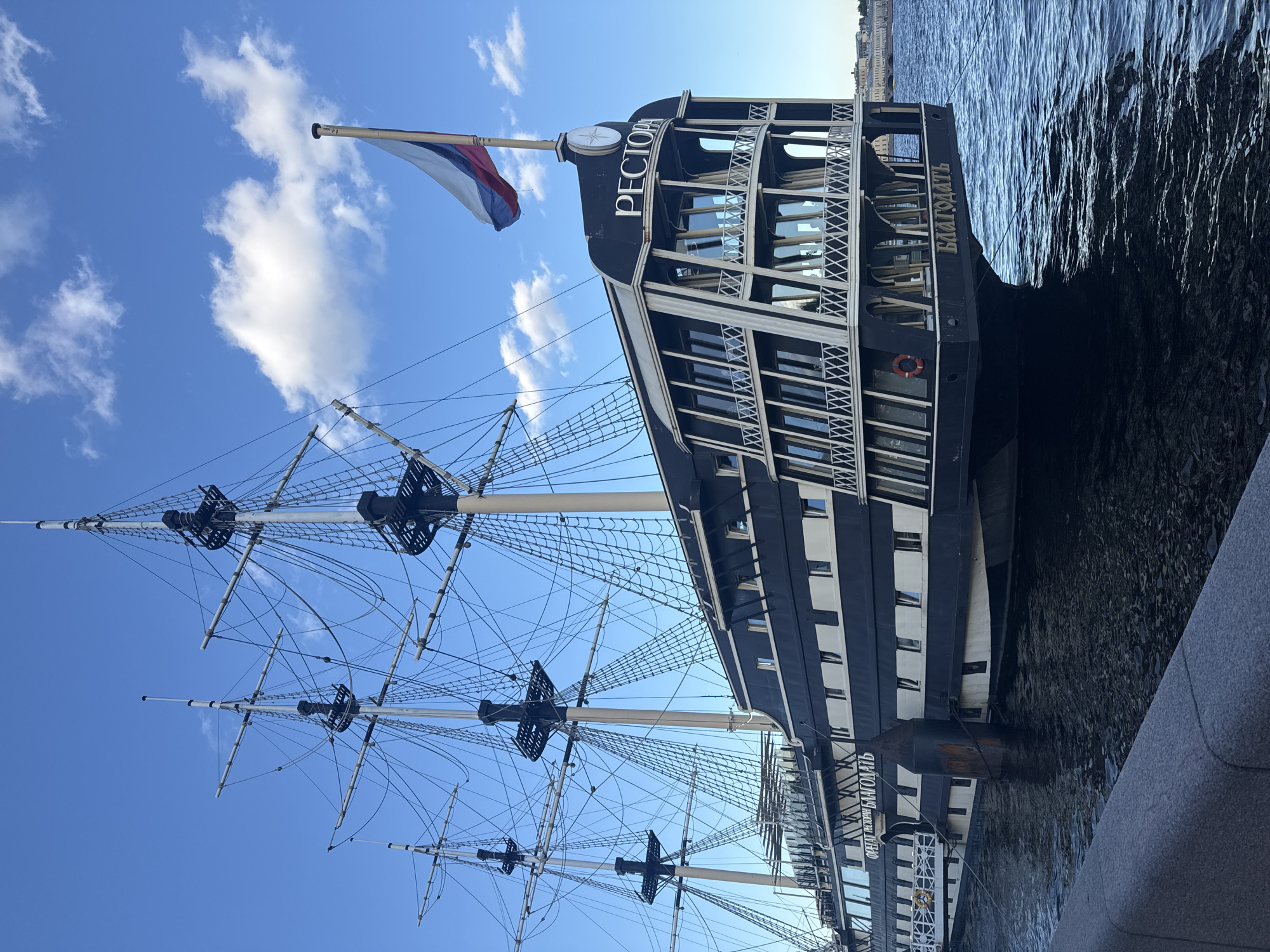 A restored tall ship, the "Blagodat" (Благодать), is moored in the Neva River in Saint Petersburg, Russia. The ship, with its three masts and intricate rigging, is painted black and white and functions as a restaurant. The Russian flag flies from the stern. The sky is blue with scattered white clouds. The water is dark and choppy. A stone embankment runs along the foreground. The ship's name, "Blagodat," is visible on the stern in gold lettering. The word "Ресторан" (Restaurant) is also displayed on the stern. The ship appears to be a tourist attraction.