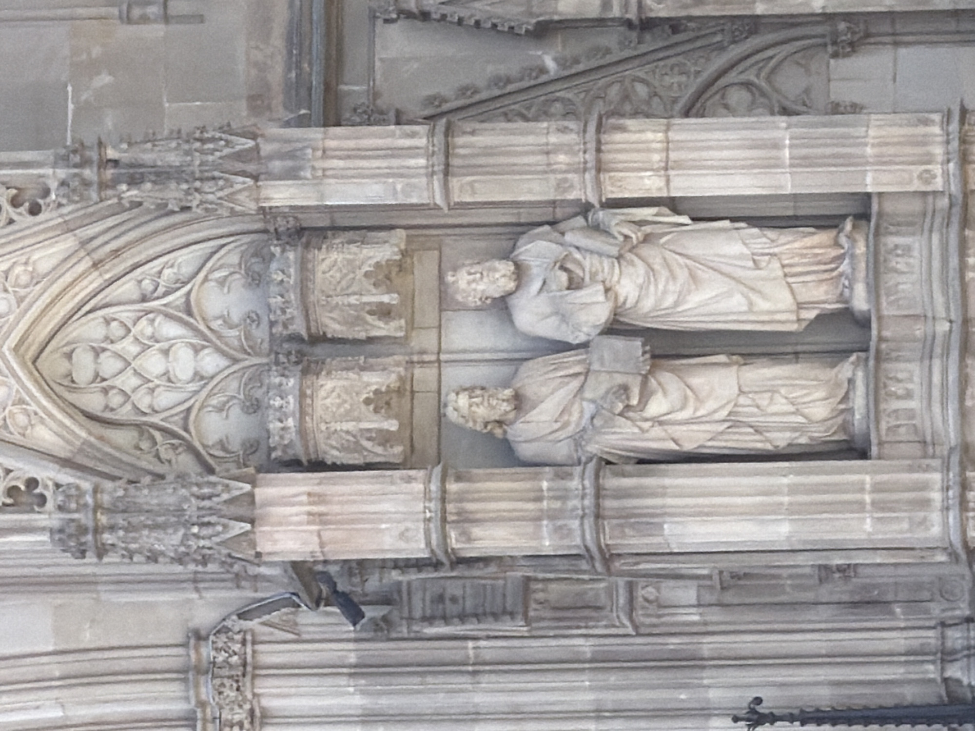 The image shows two stone statues of male figures standing in niches on the exterior of a building. The figures are dressed in robes and hold books. They appear to be saints or apostles. The architectural style is Gothic, with pointed arches and intricate stonework. The statues are weathered and appear to be very old. The building is made of light-colored stone.
The location is the Barcelona Cathedral, also known as the Cathedral of the Holy Cross and Saint Eulalia, in Barcelona, Spain. The statues are located on the exterior of the building, likely part of the facade or a side chapel.