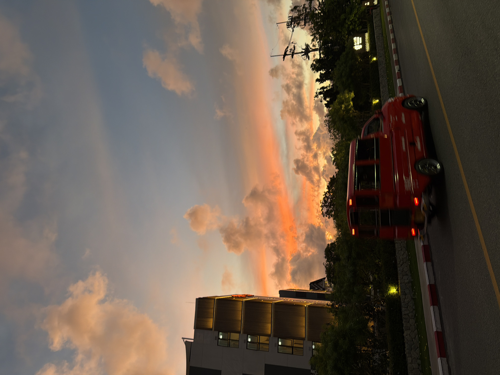 This is a vertical, eye-level, outdoor, daytime shot of a street scene in Choeng Thale, Thailand. The image captures a vibrant sunset sky with clouds tinged with orange and pink. In the foreground, a bright red bus, likely a tour bus or public transport vehicle, is driving on the right side of a paved road. The bus has multiple windows and its red brake lights are illuminated. The road is marked with a yellow line.
To the left of the road, there's a sidewalk with red and white striped borders, followed by a stone retaining wall and a strip of grass. Beyond the grass, there are trees and a modern building with illuminated windows. The building appears to have a metallic, ribbed facade and is lit from within, suggesting it might be a hotel or office building. Electrical poles and wires are visible against the colorful sky, indicating the presence of infrastructure.
The scene evokes a sense of transition as day turns into night, with the dramatic sunset colors creating a beautiful backdrop. The overall mood is calm and scenic, highlighting the natural beauty of the sky juxtaposed with man-made elements. No visible text is present in the image.