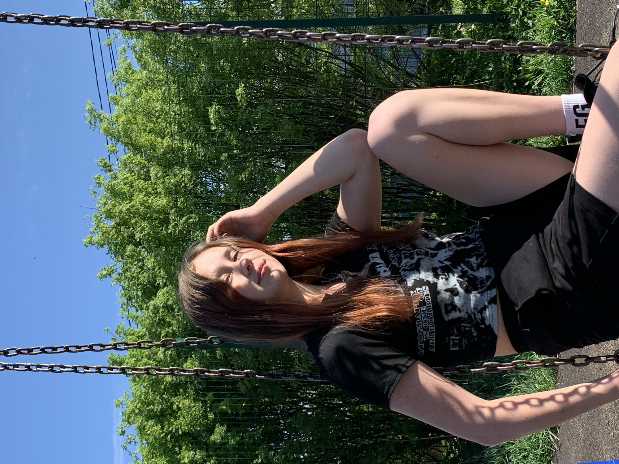 A young woman sits on a swing, possibly in a park, under a clear blue sky and a lush green canopy. She has reddish-brown hair, wears a black graphic t-shirt, and black shorts. Her legs are crossed. The location cannot be determined as Фоки, Russia.