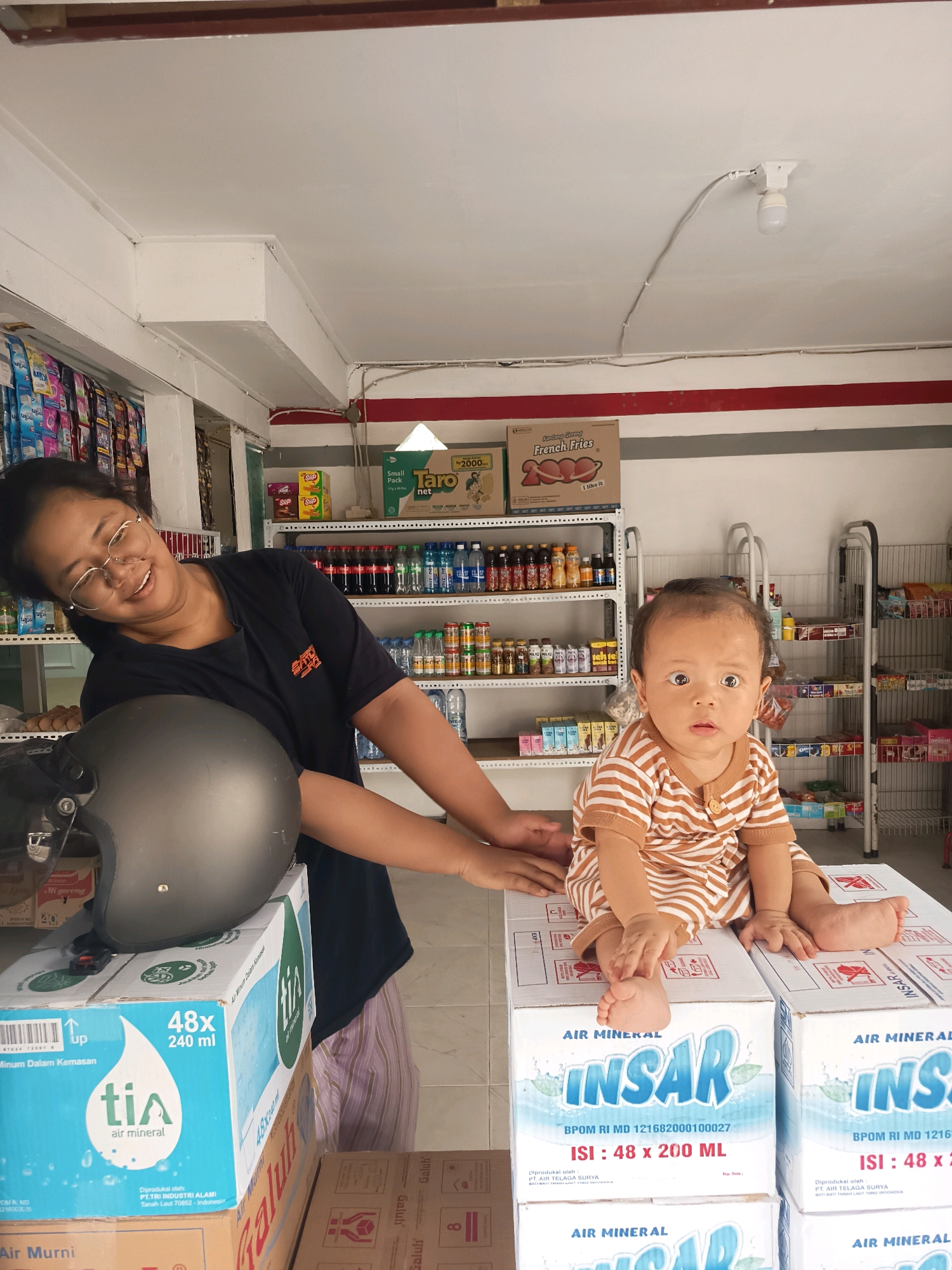 A woman with glasses, wearing a black t-shirt and striped pants, is in a small convenience store, leaning over to touch a grey helmet. Next to her, a baby in a striped onesie is sitting on a stack of bottled water boxes. The store is stocked with various products on shelves. The location is identified as Banjarmasin, Indonesia.