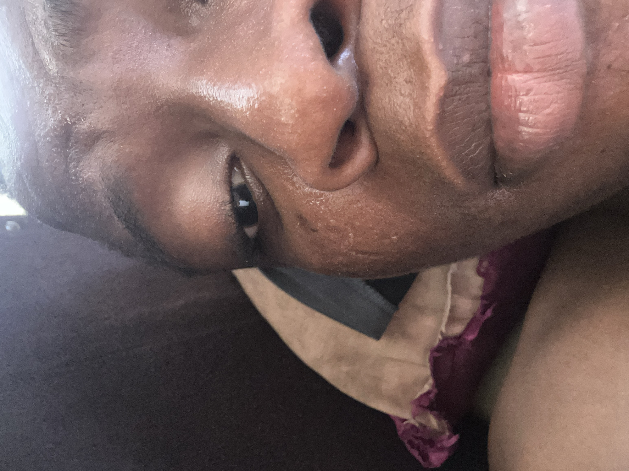 A close-up, tilted portrait of a person with dark skin. The focus is on the upper portion of their face, showcasing their nose, eye, and mouth. Their skin appears shiny, possibly due to perspiration. The individual seems to be wearing clothing, with a small portion of a patterned fabric visible near their shoulder. The background is indistinct. The location is Unknown, Unknown.