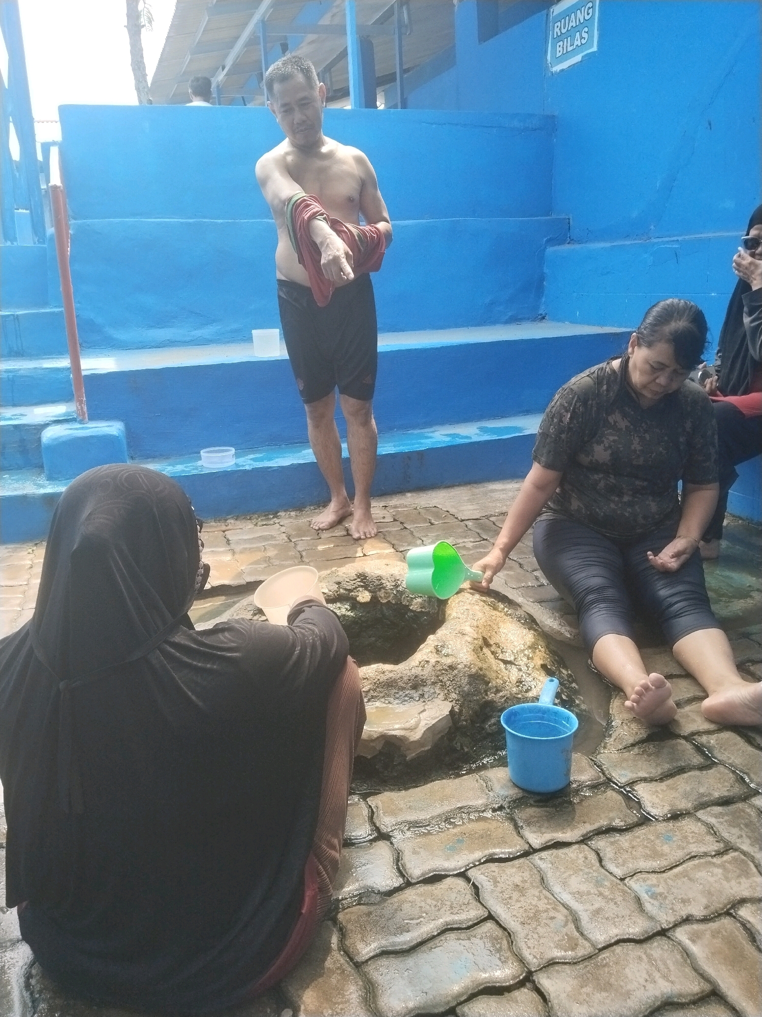 In the city of Batukuwung, Indonesia, at a geothermal spring, three individuals are visible. In the background, a shirtless man, wearing black shorts, stands near a blue wall with stairs. He is holding a red towel. In the mid-ground, a woman with dark hair is seated on the wet ground, wearing a dark patterned shirt and dark leggings. She is holding a green ladle and looking down. In the foreground, another person, wearing a black hijab and dark clothing, is also seated on the wet ground, facing away from the camera. They are holding a white bowl. A blue bucket and a natural rock formation are also present in the scene. The ground is paved with interlocking stones, and there is water on the ground.