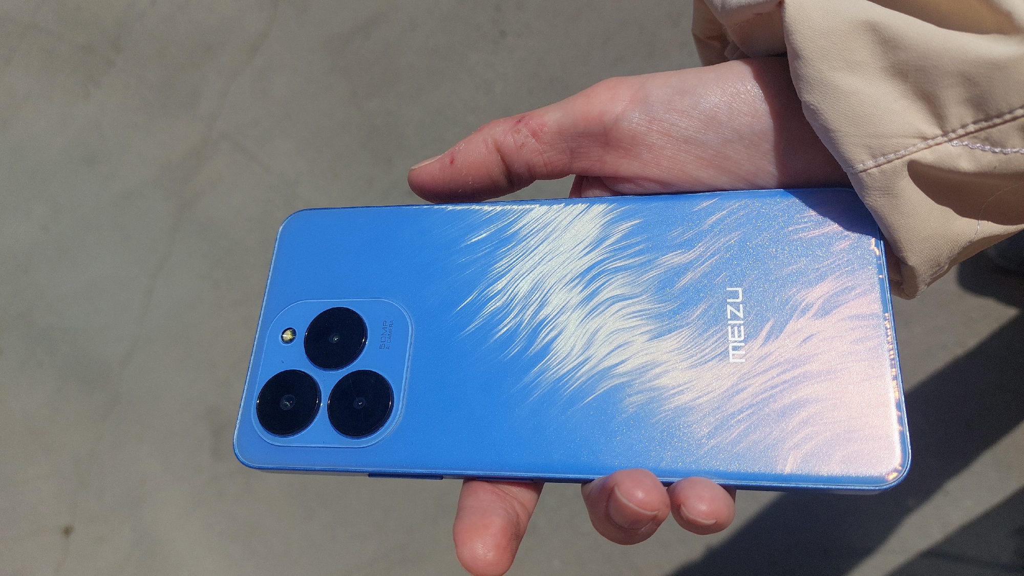 A person is holding a blue Meizu smartphone in their right hand, likely outdoors on a sunny day. The phone has three visible camera lenses arranged in a triangular formation near one end. The brand name "MEIZU" is printed on the back of the phone. The phone surface has a pearlescent effect that gives it a marbled appearance. The environment appears to be an urban or suburban setting, likely in Novosibirsk, Russia. The exact location within the city cannot be ascertained from this image alone.