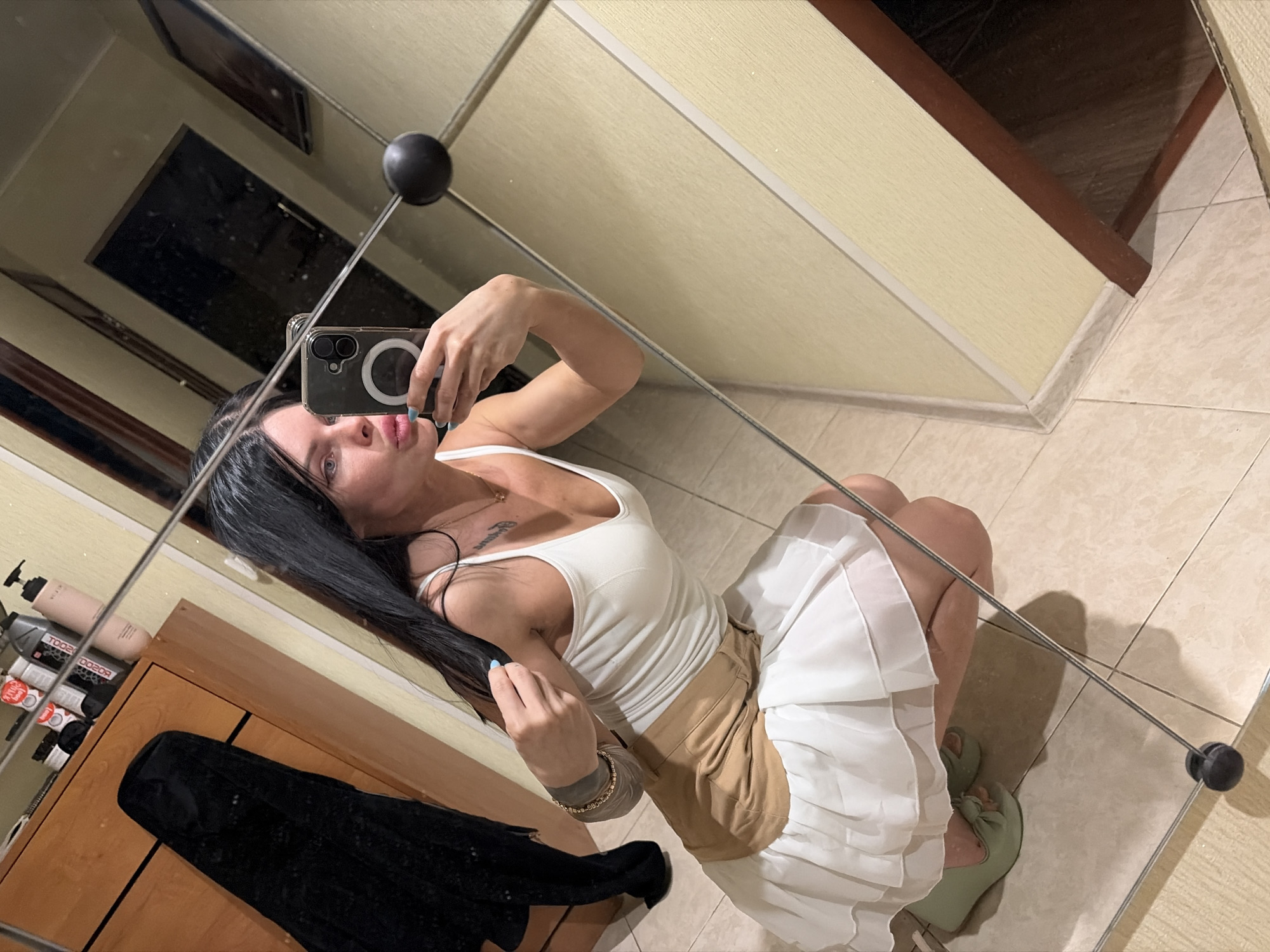 A woman with long dark hair is reflected in a mirror, taking a selfie. She is wearing a white tank top, a tan skirt, and a white ruffled skirt. Her right hand is holding a smartphone, and her left hand is touching her hair. She is crouching on a tiled floor in what appears to be a room with wooden furniture and toiletries on a shelf. The reflection also shows a black garment hanging on the furniture, a closed door, and a framed picture on the wall. The environment suggests a domestic setting.