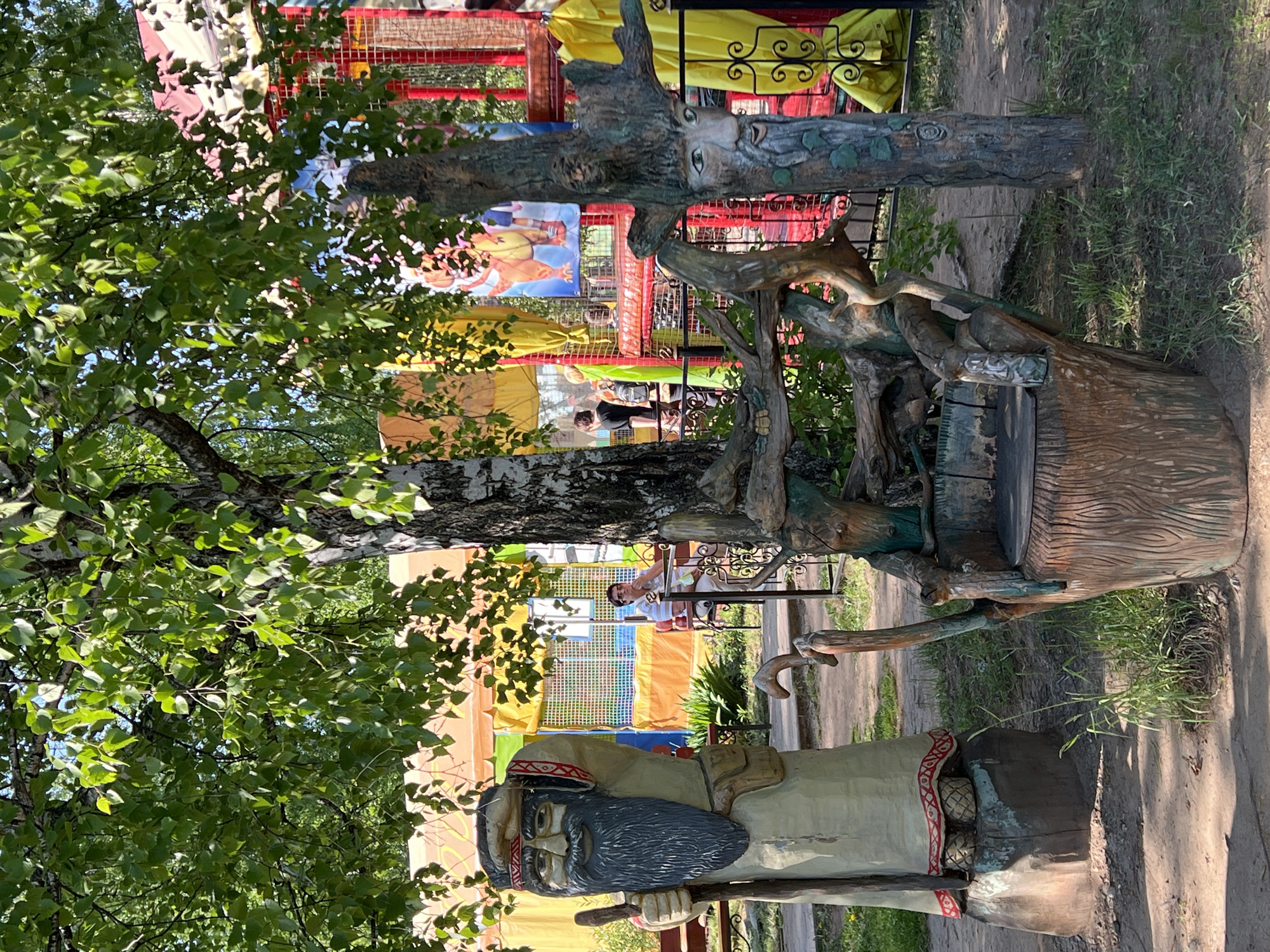 This is an outdoor image of the "Lukomorye" park in Chaikovsky, Perm Krai, Russia. The park features fairytale-themed sculptures and attractions.
The central subject is a wooden sculpture of a wizard-like figure with a long beard, dressed in traditional clothing, holding a staff. Adjacent to the figure is a wooden structure resembling a tree trunk with crafted wooden branches and a seat. At the top of the structure is another sculpted piece of wood with a face carved into it.
In the background, partially obscured by the tree, is a colorful amusement structure with yellow and red elements and hanging posters. A woman is seated within this structure. A tree with green leaves partially obscures the scene.
The ground is a mix of dirt path and grass. The scene is in daylight, with shadows indicating a sunny day.