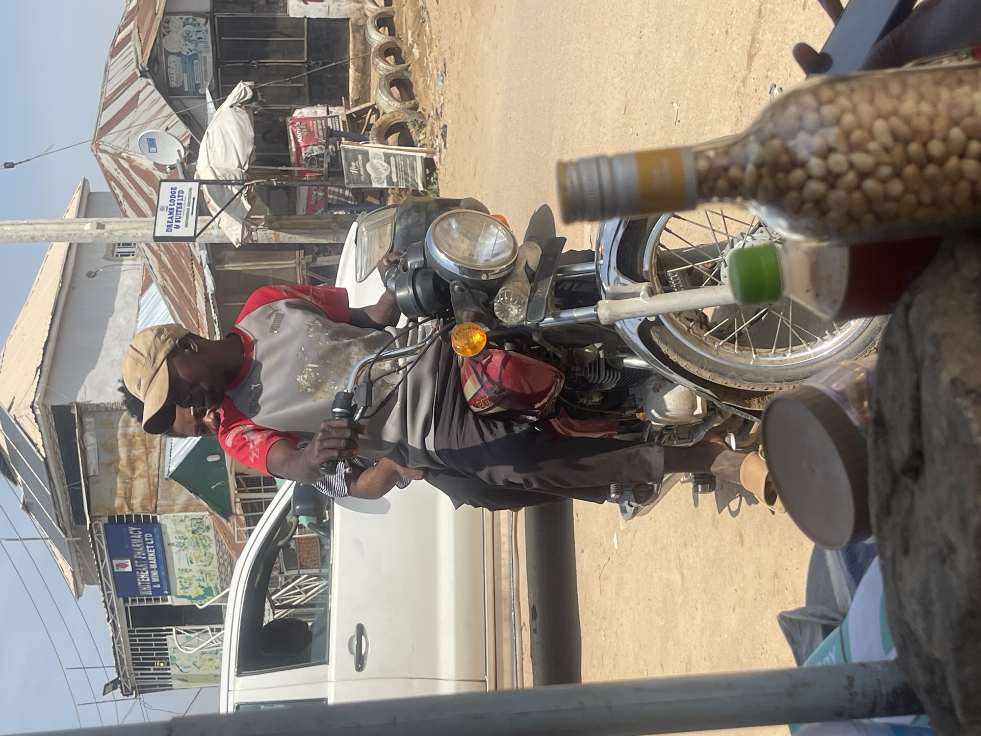 The image depicts a street scene in Abuja, Nigeria, with a man sitting on a motorcycle. The man is wearing a cap, a red and grey t-shirt, and black pants. He is holding onto the handlebars of the motorcycle. In the foreground, a glass bottle filled with peanuts is visible, placed in front of the motorcycle's wheel.
The background reveals a bustling street with various shops. Signs for "DREAMS LODGE & SUITES LTD" and "WHITEHEART PHARMACY & MINI-MARKET LTD" are prominent, suggesting a commercial area. Other visible text includes "PAULA'S," "Annie's," and "New CALABAR KITCHEN." Several tires are stacked along the side of the street, and the ground appears to be dusty, indicating a dry environment. The sunlight suggests it is daytime. The overall atmosphere appears to be that of a typical day in a Nigerian town or city.