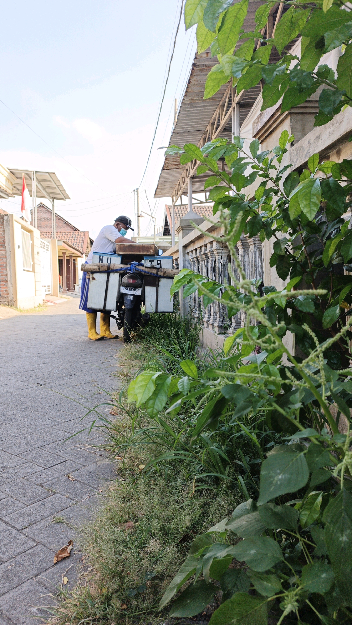 In the city of Ketegan, Indonesia, a person wearing a white shirt, a blue and white skirt, yellow boots, and a face mask stands next to a motorcycle. The motorcycle is equipped with two large white boxes attached to the back, and a wooden board on top. The person appears to be interacting with the boxes on the motorcycle. The scene is on a paved pathway next to a building with a decorative fence. Green foliage is prominent in the foreground, obscuring part of the building. The background shows more buildings, a red and white flag, and overhead wires. The sky is clear and bright.