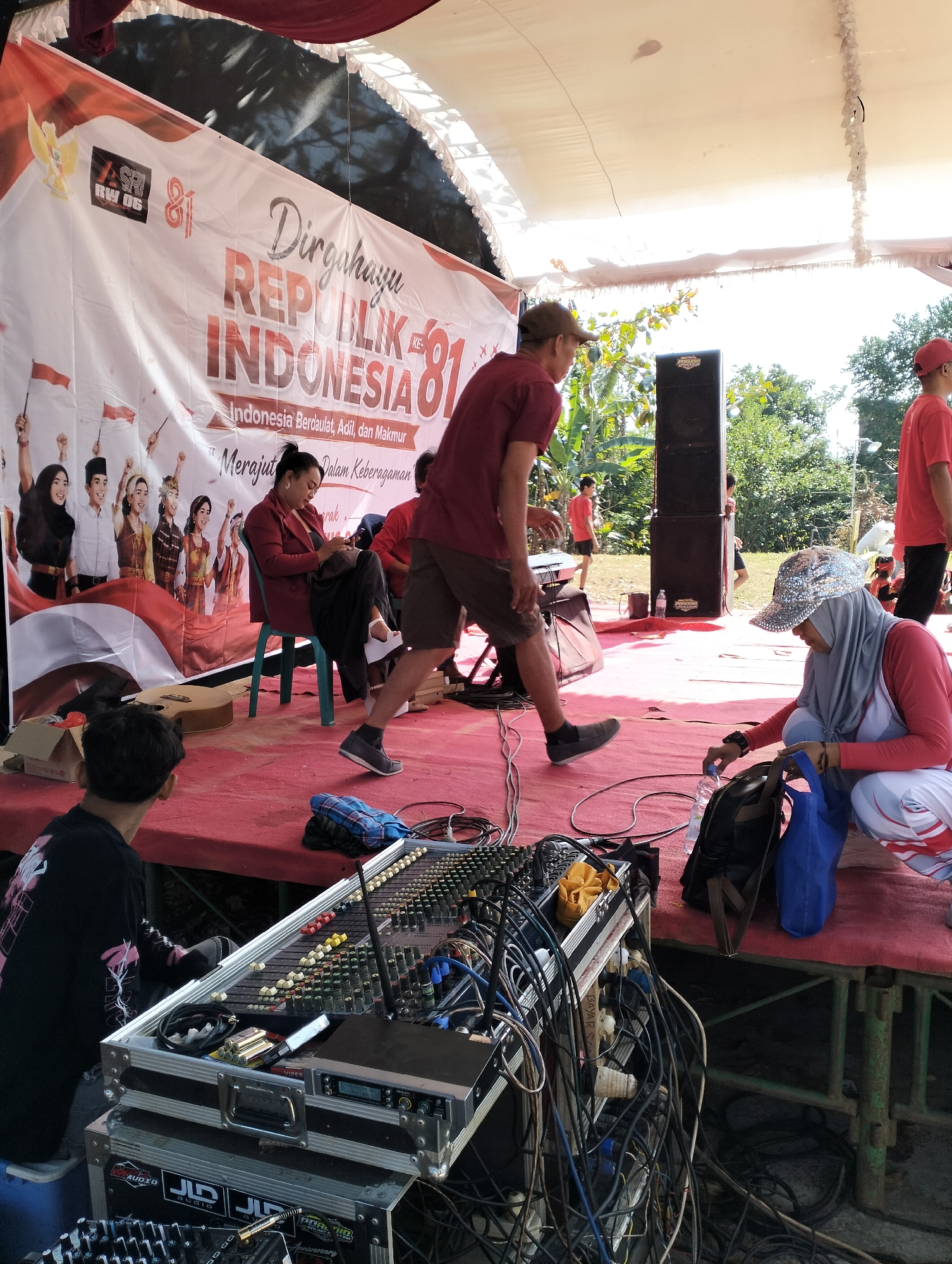 The image captures an outdoor event, likely a stage setup for a celebration, on a sunny day. A large banner in the background reads "Dirgahayu REPUBLIK INDONESIA KE-81," indicating a celebration of Indonesia's 81st Independence Day. The stage is covered with a red carpet, and a sound mixing board with numerous cables is prominently placed in the foreground. Several individuals are present on or near the stage.
In the foreground, a person wearing a black long-sleeved shirt sits near the sound equipment. To the right, a woman wearing a grey hijab and a sparkly cap is crouched down, holding a water bottle and a blue bag. She is wearing a pink and white outfit. Further back on the stage, a woman in a red blazer is seated on a turquoise chair, looking down at her lap. Another person in a maroon t-shirt and shorts is walking across the stage, and another in a red t-shirt is standing in the background. Various speakers and equipment are set up on the stage, with lush green foliage and trees visible behind the stage. The overall scene suggests preparation or a break during a live event. The image does not provide specific visual cues to pinpoint the exact location in Semarang, Indonesia, but it is clearly an outdoor public gathering.