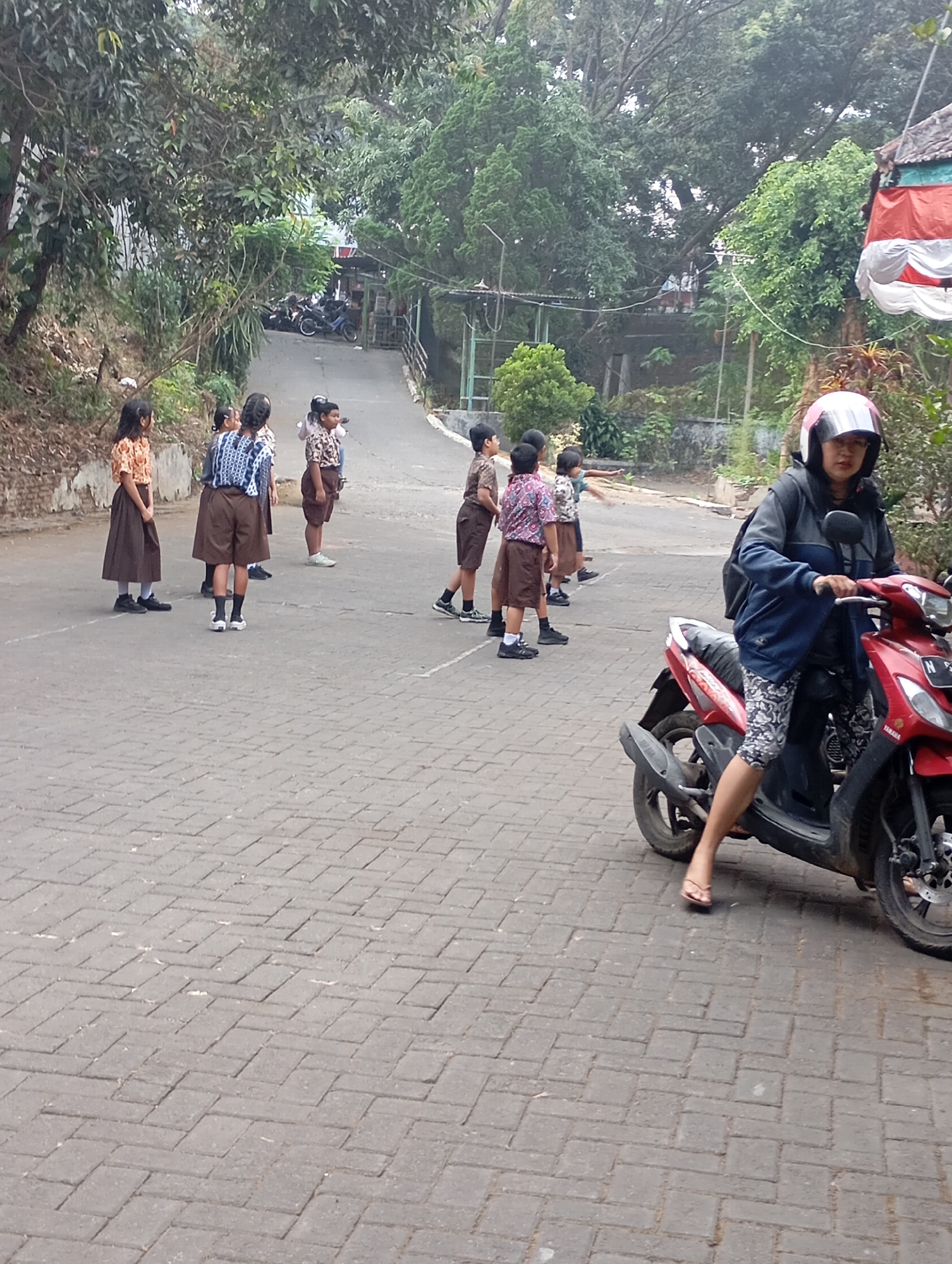 In Malang, Indonesia, a group of schoolchildren in uniform are standing on a paved road surrounded by trees and buildings. A woman wearing a helmet and a jacket is riding a red motorcycle in the foreground. The children appear to be waiting for something, with some looking in the direction of the motorcycle and others looking elsewhere. The road is paved with interlocking bricks and slopes upwards into the distance. Buildings are visible in the background, along with parked motorcycles. The overall scene suggests a moment captured during a typical day in a school environment in Malang.
