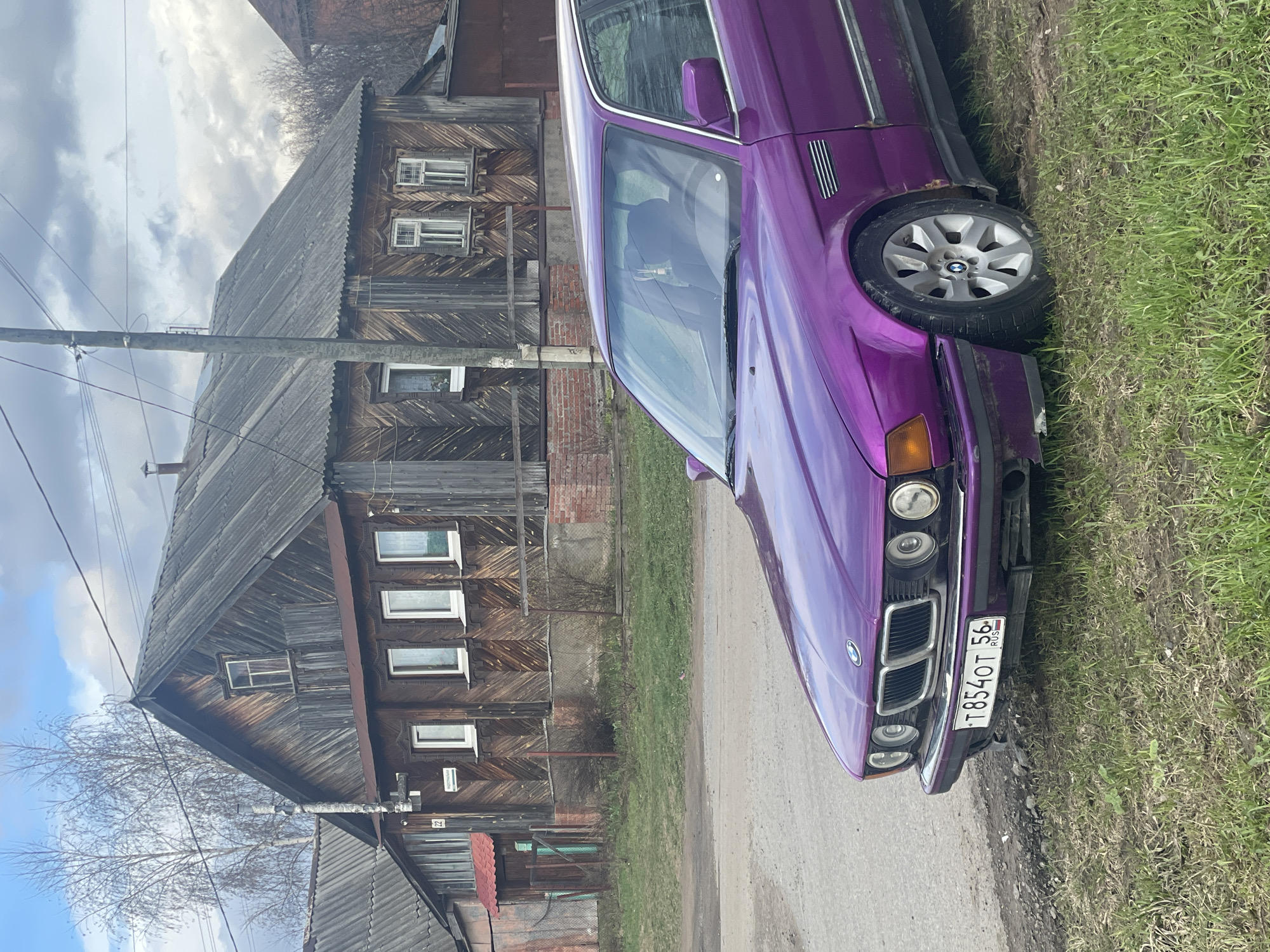 The image, rotated 90 degrees clockwise, depicts a vibrant purple car parked on an unpaved road next to a traditional wooden house under a partly cloudy sky.
The central subject is a glossy purple BMW sedan, an older model identifiable by its distinctive front grille and emblem on the hood. The car is equipped with multi-spoke silver alloy wheels. A Russian license plate is clearly visible on the front bumper, displaying the characters "T854OT 56 RUS". The vehicle shows some signs of wear on its front bumper and around the wheel arches.
The car is situated on a dirt or gravel road, with a strip of green grass and some muddy patches separating it from the weathered, two-story wooden house in the background. The house features a dark, horizontally paneled wooden facade and numerous white-framed windows, some with decorative surrounds. It is topped with a grey, corrugated roof, and a brick chimney is visible. A wooden utility pole with power lines stands directly in front of the house, partially obscuring the view. Bare deciduous trees, suggesting early spring, are visible against the bright, overcast sky. A small sign with the number "21" is affixed to the house near a lower window. The scene evokes a sense of a quiet, possibly rural or older semi-urban setting, consistent with the described location context of Izhevsk, Russia.