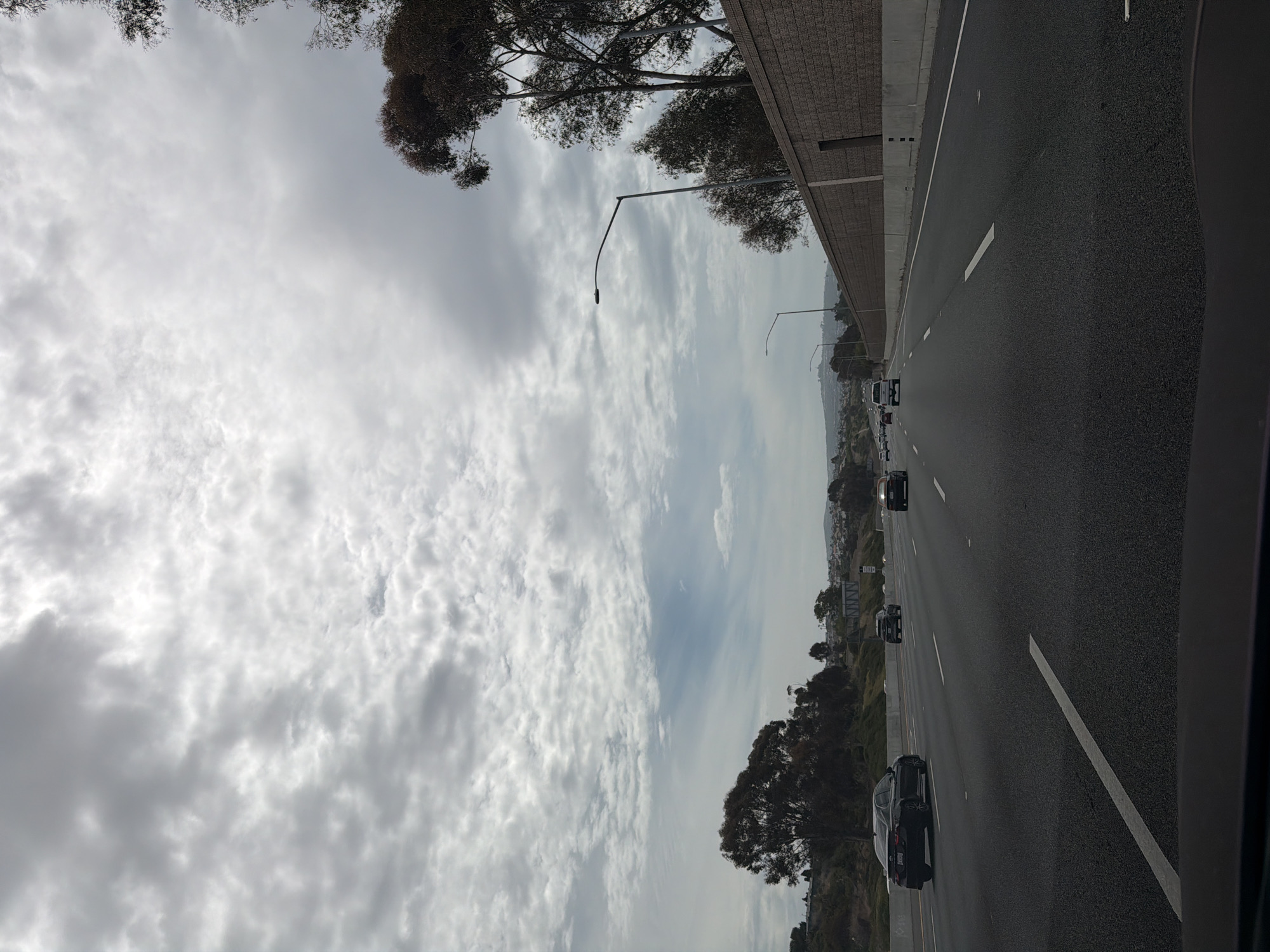 The image captures a view from a vehicle traveling on a multi-lane highway. The sky is overcast with dense, textured clouds. On the right side of the highway, a textured brick retaining wall runs alongside the road, with trees and vegetation visible beyond it. On the left side, there are rolling hills with scattered trees and buildings in the distance. Several cars are visible on the highway, all moving in the same direction. The road itself is asphalt with clear white lane markings. Streetlights are present along the highway. The scene suggests a cloudy day in an urban or suburban area, possibly San Clemente, California, given the typical coastal highway architecture and vegetation.