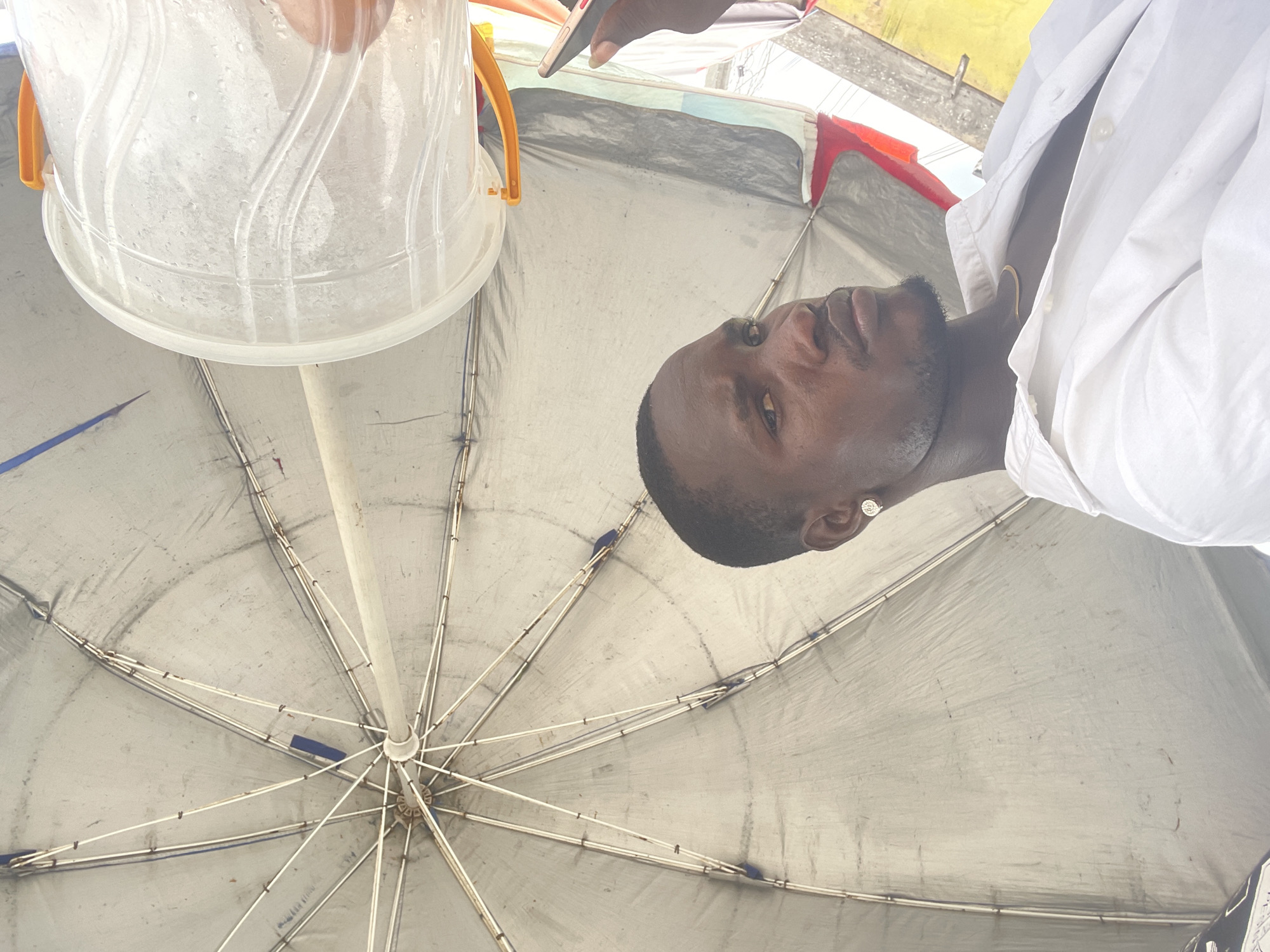The image shows a low-angle, point-of-view shot looking upwards under a large, gray umbrella. A person is visible partially within the frame, wearing a white collared shirt. They have short, dark hair and facial hair. The person is close to the camera. Above the umbrella and to the left, a translucent bucket-like object is visible, partially filled with a substance that appears to be granular. Above the umbrella at top center, a hand is holding what appears to be a smartphone. The umbrella’s support structure is visible. The environment is suggestive of an outdoor setting in Ado, Nigeria, though specific location details are not discernible. The action observed suggests a selfie or a low-angle photograph is being taken.