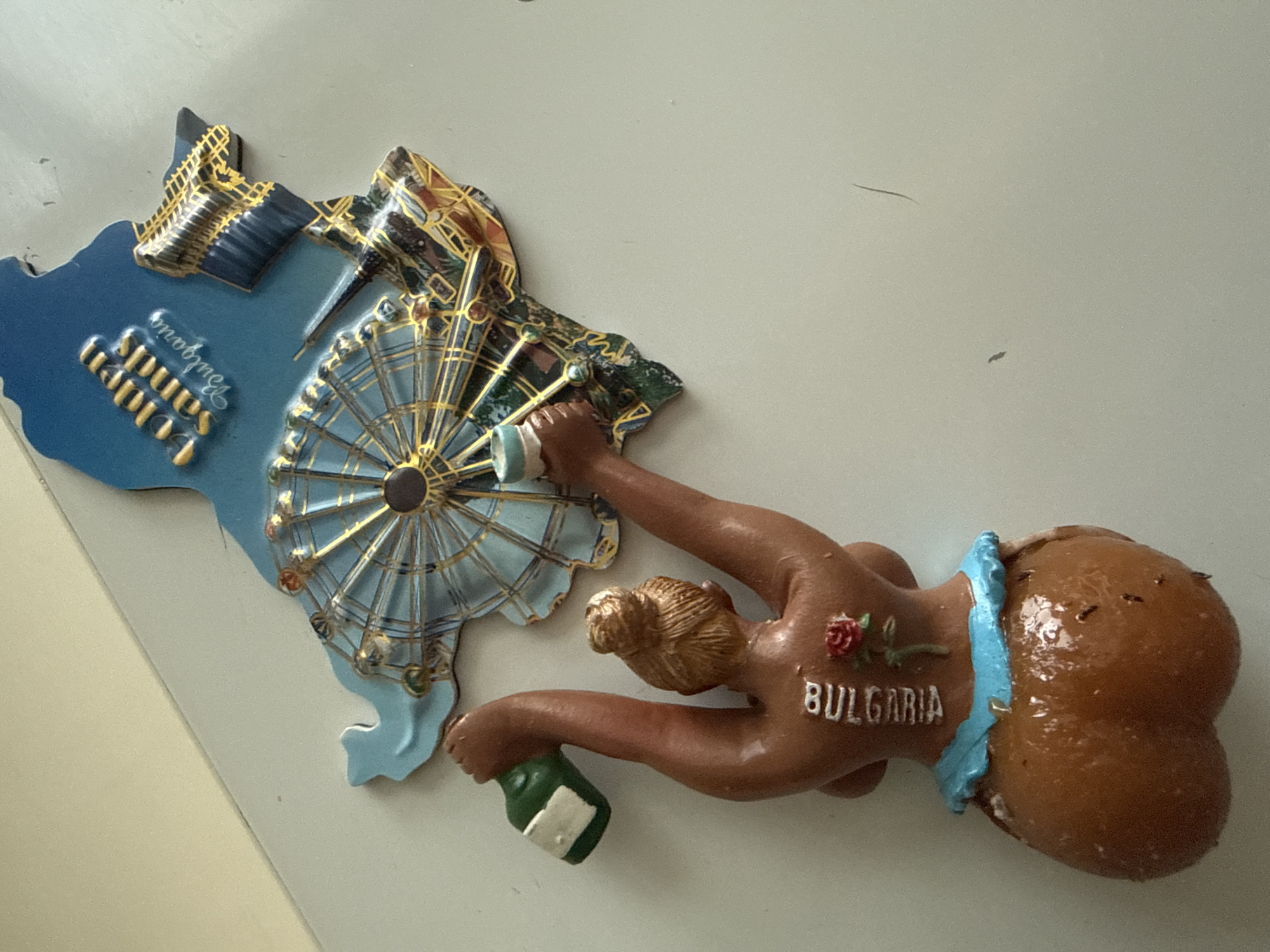 The image shows a souvenir magnet depicting a female figure and a map-shaped representation of a location. The female figure is brown-skinned with blonde hair in a bun. She is holding a green bottle in her left hand and a cup in her right hand. The word "BULGARIA" is written vertically on her back, above a red rose tattoo. The bottom of the figure appears to be a stylized representation of buttocks, covered with a blue garment.
The map-shaped part of the magnet is blue and appears to represent a coastal area, possibly a beach resort. Text on the map reads "Golden Sands Bulgaria". It features a Ferris wheel and what looks like buildings or a monument. The Ferris wheel is gold and blue, with colorful gondolas.
The souvenir is affixed to a light-colored, flat surface, likely a refrigerator door. The surface shows minor scuff marks and is slightly textured. The lighting suggests an indoor environment with overhead illumination. There are no people or animals present besides the figurine. The image is taken from a slightly elevated angle, looking down at the magnet. The background is out of focus, showing a light-colored wall or surface with some minor imperfections. The image does not contain any visual elements that would indicate the location is the city of Cahul, Moldova.