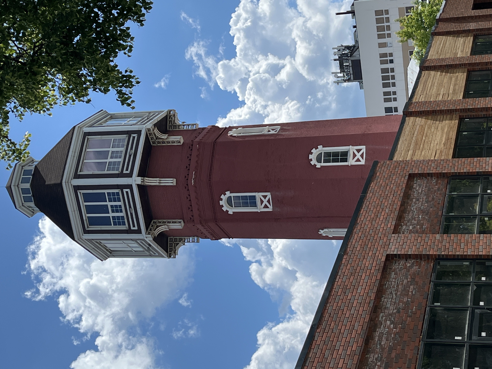 The image, rotated 90 degrees clockwise, prominently features a tall red brick tower with a dark, possibly conical, roof. The upper section of the tower is adorned with ornate white trim, framing several multi-paned dormer windows. Further down, smaller arched windows with white frames and decorative cross patterns are visible on the brick facade.
To the right of the tower, a modern building with a facade of light brown brick and horizontal wooden panels is partially visible, featuring large, dark-paned windows. In the background, another white building with numerous small, square windows and communication antennas on its roof can be seen. A lush green tree branch extends into the upper left corner of the frame.
The bright blue sky and prominent white clouds indicate a sunny daytime setting. There are no people visible, nor is any specific activity taking place. The scene suggests an urban or semi-urban environment, showcasing a blend of architectural styles. No discernible text is present in the image.