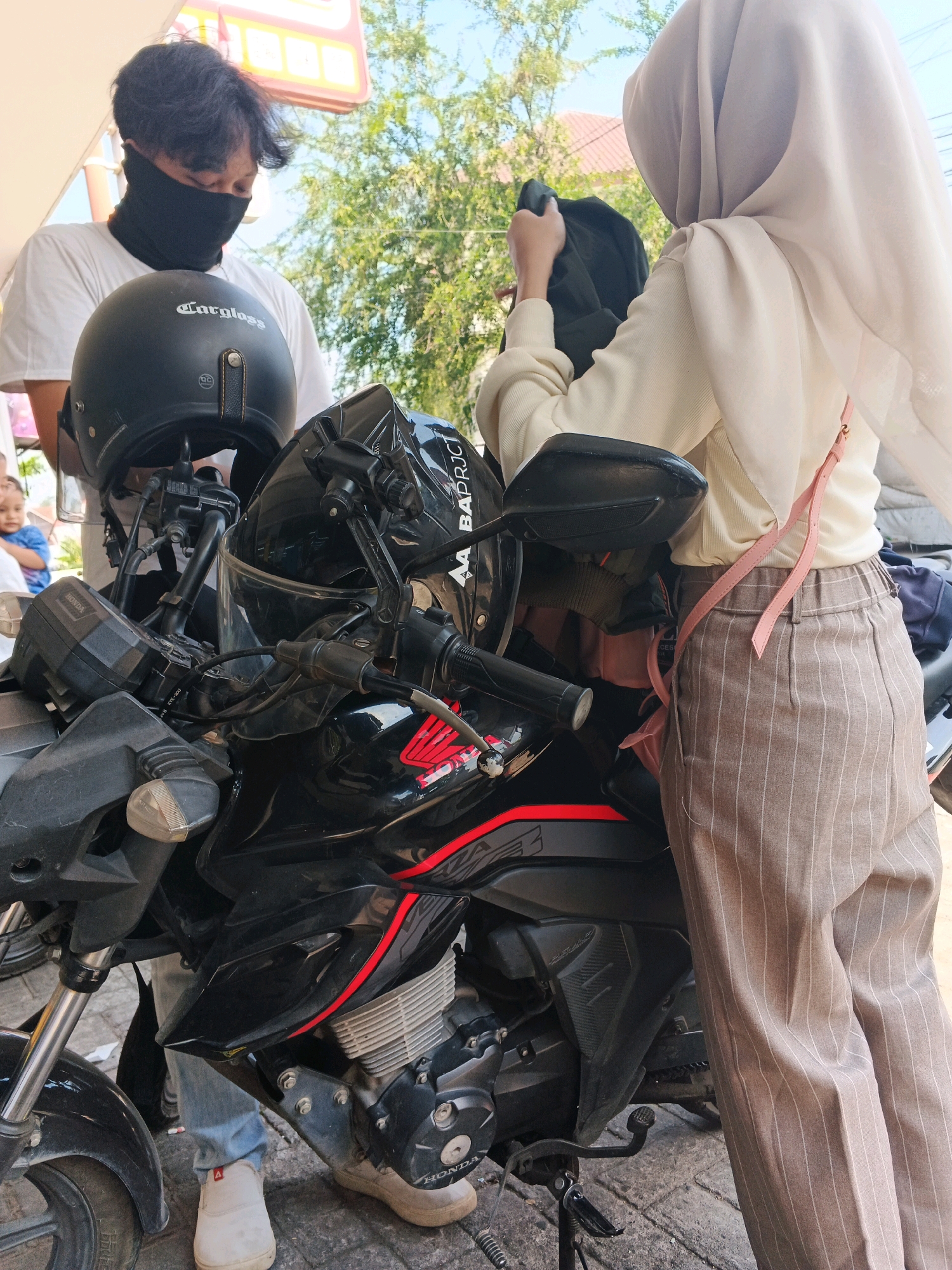In Kalirejo, Indonesia, two individuals stand near a black motorcycle. The person on the left, identified as male based on attire and hairstyle, wears a white t-shirt and a black neck gaiter covering their lower face. A black helmet with the "Cargloss" logo rests on the motorcycle in front of them. The person on the right, identified as female due to the hijab and clothing, wears a cream-colored top and striped brown trousers. They are holding a dark garment. The motorcycle is equipped with a helmet mount on the handlebars and appears to be parked on a paved surface, possibly a sidewalk or road. The background shows greenery and hints of buildings, consistent with an urban or semi-urban setting.