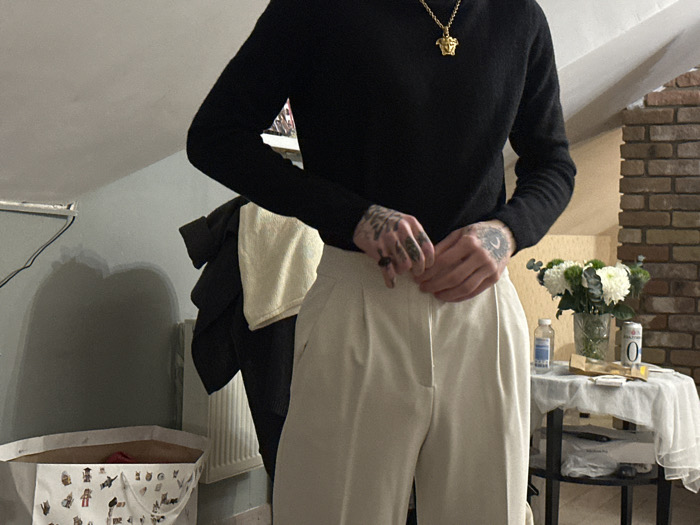 A person wearing a black long-sleeved shirt and white trousers fastens their pants. They wear a gold necklace. A clothes rack with a black coat and cream-colored towel stands to the left. To the right, a small table covered with a white cloth holds a vase of white flowers, a bottle, a small box, and a can. A brick chimney is visible behind the table. In the background, the room has angled walls and a laundry basket with drawings on it.