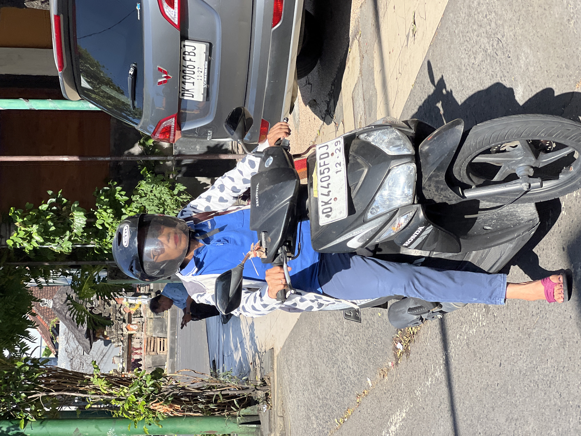 In Kuta, Indonesia, a person wearing a black helmet with a clear visor and a blue polo shirt over a white and grey patterned jacket rides a black scooter. The scooter has a license plate that reads "DK 4405 FDJ" and "12.29". The rider is wearing blue pants and pink sandals. A grey car with a license plate "DK 1906 FBJ" is parked to the right of the scooter, and its emblem appears to be a red "W". The background shows a street scene with buildings, trees, and foliage. Another person is visible in the background sitting on a curb and looking at a mobile phone. The sun is casting shadows on the road.
