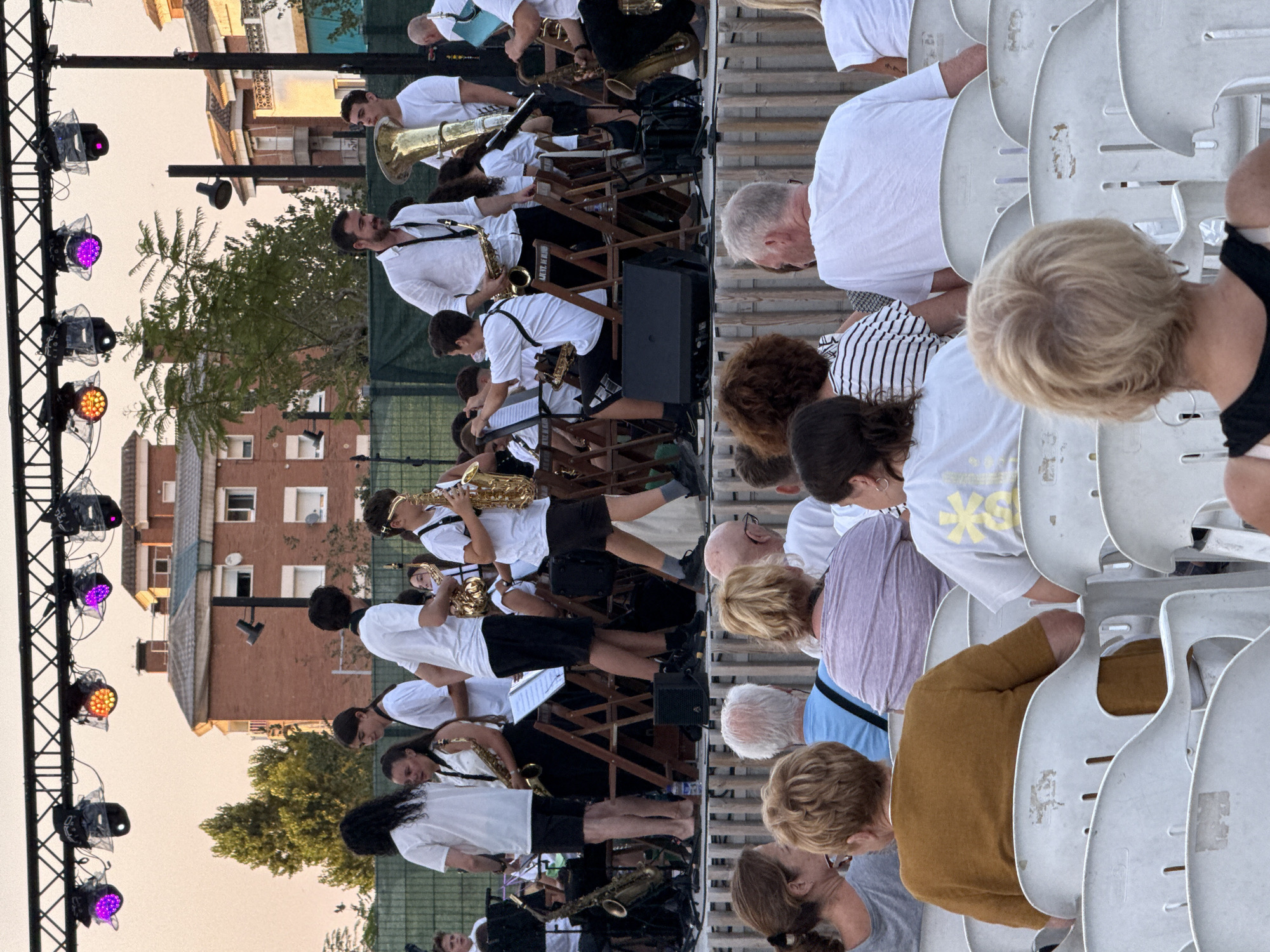This is a medium shot, eye-level, outdoor photograph of a band performing on a stage, with an audience seated in white plastic chairs in the foreground. The band members are dressed in white shirts and black shorts or pants, and many are playing instruments such as saxophones and a tuba. The stage is lit with colorful lights, and a building with red brick and white trim is visible in the background. The scene appears to be taking place in the city of Blanes, Spain. The audience members are facing the stage, and some are looking at the performers, while others are looking away. The overall atmosphere is one of a summer concert or festival.