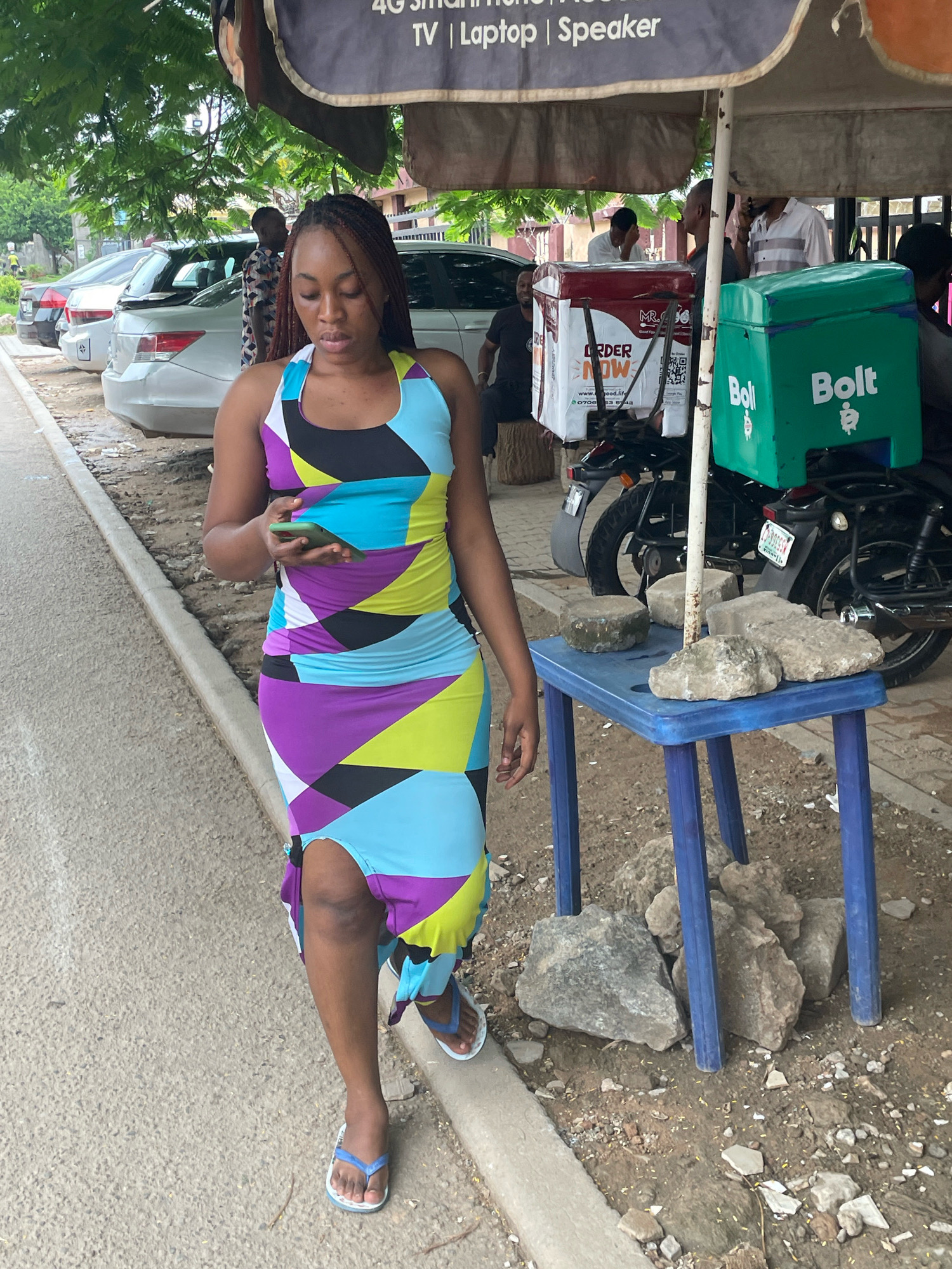 In an outdoor urban setting in Abuja, Nigeria, a young woman wearing a colorful, geometric-patterned dress and blue flip-flops walks while looking at her smartphone. She is positioned on a paved walkway next to a dirt area with scattered rocks and a blue plastic table holding more rocks. Behind her, a motorcycle is parked with two insulated delivery boxes, one red and white with "ORDER NOW" branding and the other green with "Bolt" lettering. Several people are visible in the background, some standing near vehicles and an umbrella. Trees with lush green foliage provide shade overhead. The overall scene suggests a street-side commercial area.