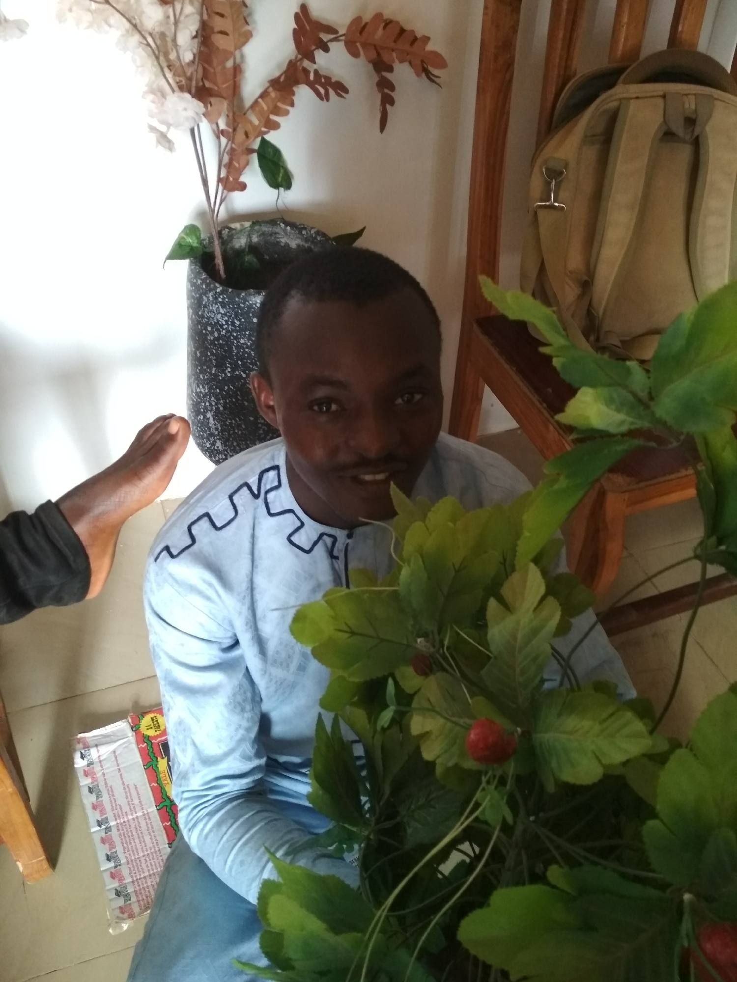 A man is squatting indoors in Unknown, Unknown. He is wearing a light blue shirt with dark blue detailing around the neck and sleeves and light blue jeans. He is partially obscured by a bush with green leaves and red fruits. A gray backpack is hanging on the wooden frame of a chair to the man’s right. A gray pot with a plant in it is to the man’s left. A foot is visible to the left of the man. There is also an open packet of tissues on the floor to the left of the man.