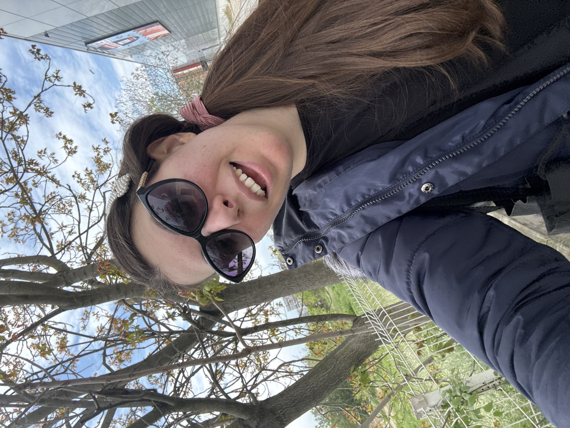 A young woman takes a selfie outdoors, likely in a city park, as implied by the presence of trees with new foliage, a metal fence, and a building in the background. She is wearing sunglasses and a dark blue puffer jacket over a black shirt, with a pink scrunchie holding back part of her long, brown hair. The image captures her smiling slightly, with a partial view of her teeth. The sky is visible between the branches of the tree, suggesting a bright day. The exact location in the city of Unknown, Unknown cannot be determined with the information available in the image.