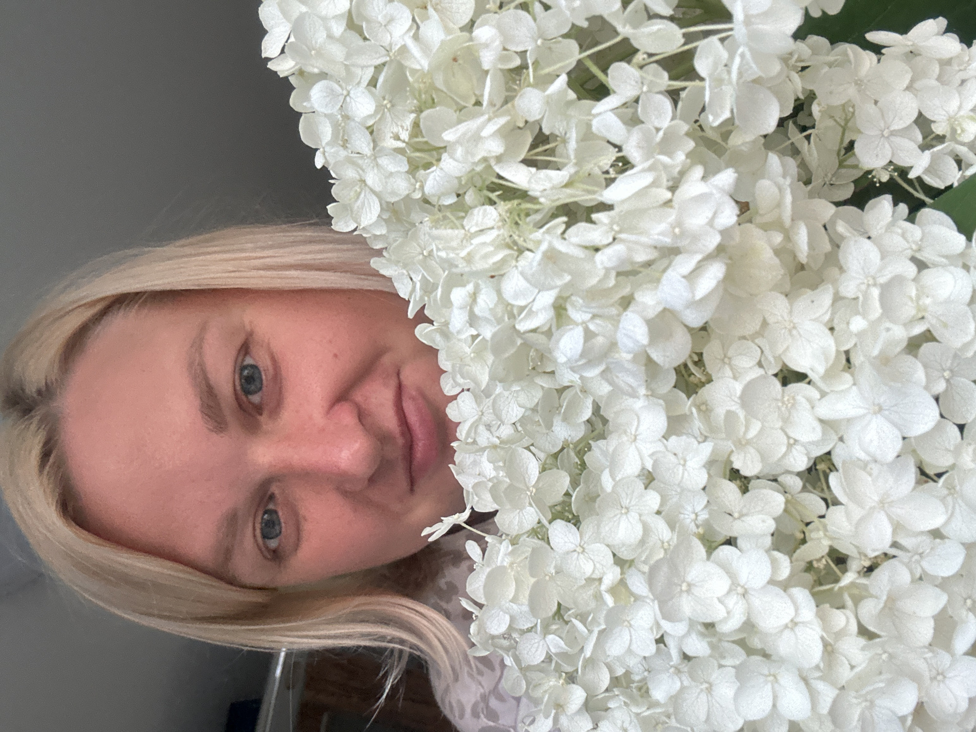 A close-up, waist-up, outdoor photograph shows a woman holding a large bouquet of white hydrangeas. The woman has fair skin, blonde hair, and blue eyes. She is smiling and looking at the camera. The hydrangeas are in full bloom and fill the foreground of the image. The background is blurred and appears to be a natural setting. There is no visible indication of the city of Chaikovsky, Russia.