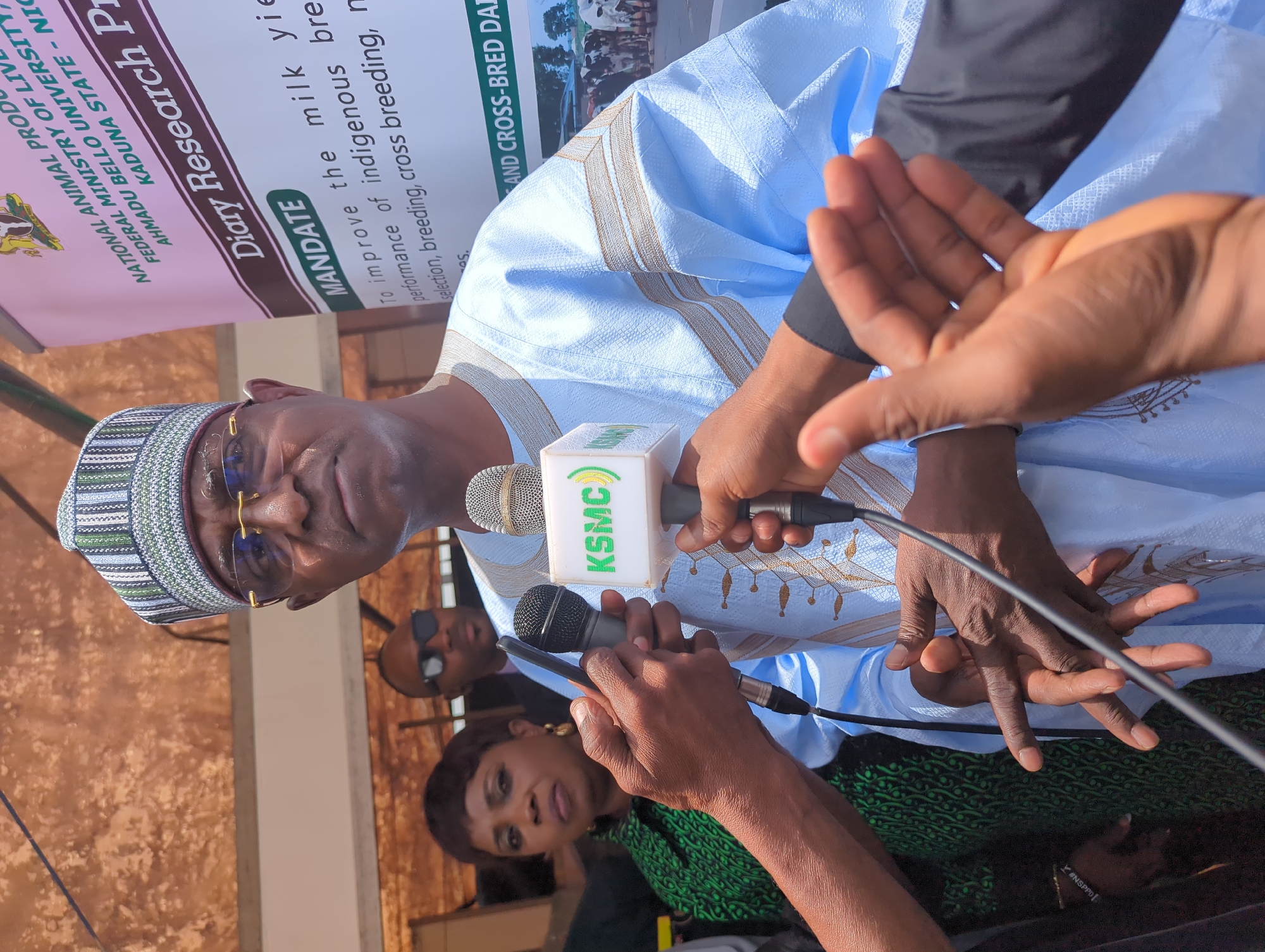 A middle-aged man in traditional African attire, including a patterned cap and glasses, is the focal point of an outdoor press conference setting. He is standing in front of a banner that reads "Diary Research Programme," which lists "MANDATE" as improving the milk yield and the performance of indigenous breeds. Microphones from various media outlets, including one with the "KSMC" logo, are positioned near his face, suggesting he is speaking or about to speak. Several reporters are holding the microphones. In the background, partially obscured figures can be seen, including a woman in a green patterned top and a man wearing sunglasses. The location details given are Unguwan Sanusi, Nigeria.