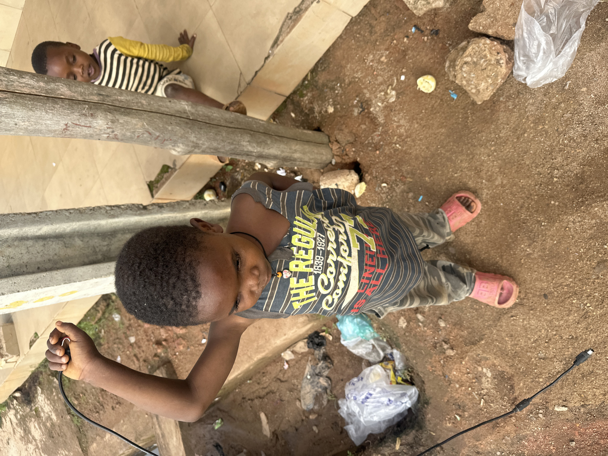 The image shows two young children outdoors, likely in an urban environment in Gura Top, Nigeria. In the foreground, a child stands looking down and to the right, holding a black cable in their raised right hand. The child is wearing a gray and black striped tank top with a graphic design that reads "THE REGUL... COMFORT" and "FINE... ALL HER", and gray pants with a black and white pattern. They are wearing pink slides.
Behind and to the right of the foreground child, another child is seated on a raised, tiled surface. This child is wearing a long-sleeved shirt with horizontal black and yellow stripes and appears to be looking to the left.
The ground around the children is dirt and sand, with scattered debris including plastic bags, rocks, and what appears to be a piece of fruit. A thick, weathered wooden post stands between the two children, partially obscuring them. A concrete pillar or structure is visible to the left of the foreground child. The overall setting suggests a casual outdoor area, possibly a residential compound or public space with some degree of disarray.