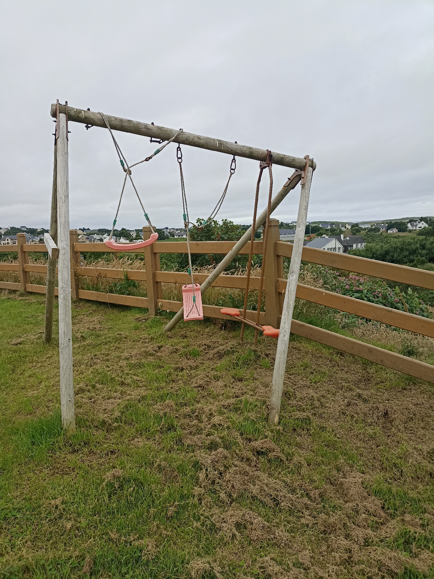 This image captures an outdoor scene with a wooden swing set as the central focus. The swing set is constructed from weathered wooden posts and a horizontal beam, from which three swings are suspended by ropes. Two of the swings are pink, one appearing to be a molded plastic seat, and the other a flat, rectangular seat. The third swing has an orange plastic seat. The swing set is situated on a grassy area with some patches of dry, matted grass. Behind the swing set, a wooden fence runs horizontally across the frame. Beyond the fence, a hillside dotted with houses and trees is visible under a cloudy sky. The overall setting suggests a rural or semi-rural environment. The image appears to have been taken in Doirí Beaga, Ireland, judging by the architectural style of the houses and the landscape. There are no people or animals visible in the image.