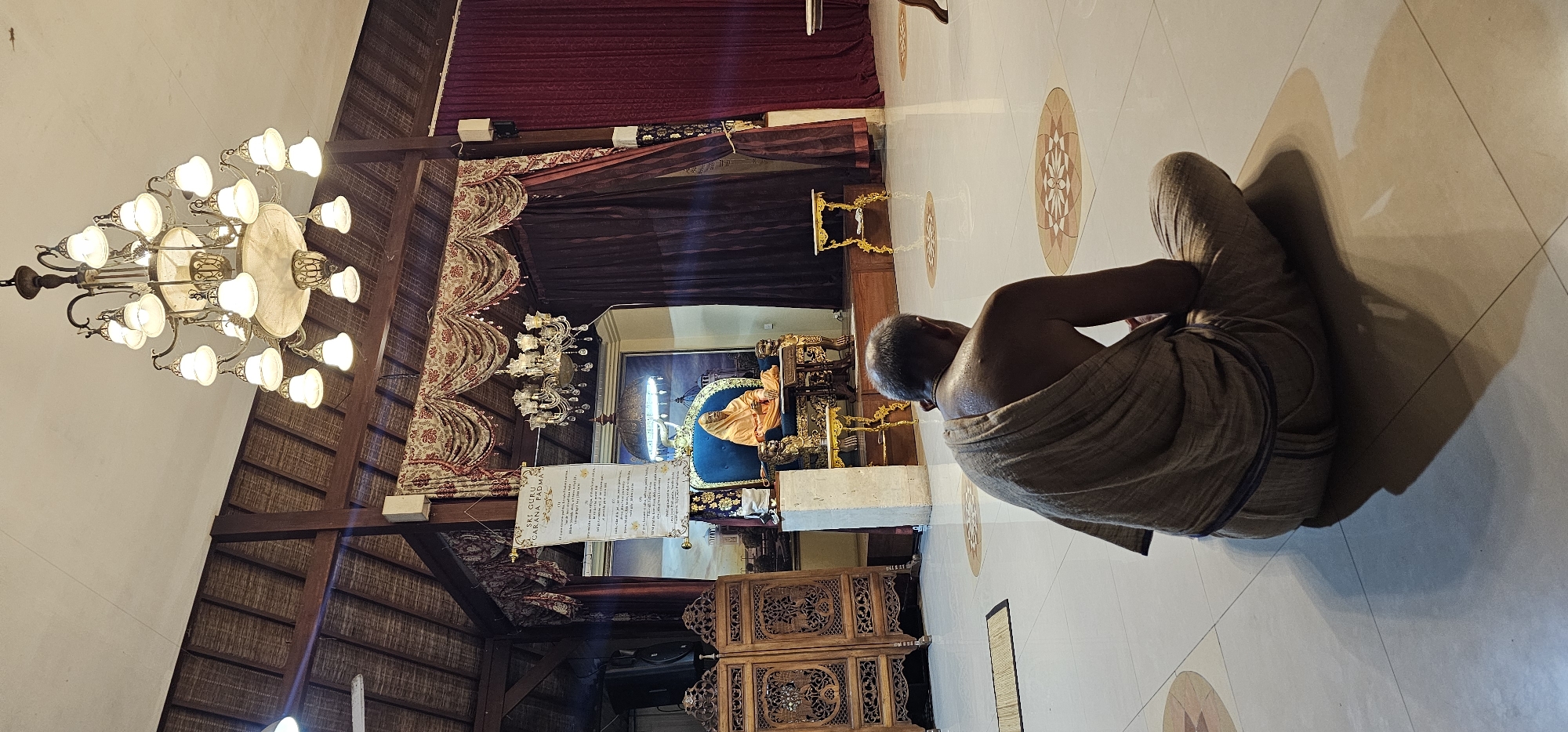 The image shows the interior of a temple, likely in Denpasar, Indonesia, based on the architectural style and objects within the space. An elderly man is seated on the tiled floor, wearing a cloth draped over his shoulders. He faces an ornate shrine or altar adorned with statues and religious artifacts, set against a backdrop of rich fabrics and decorative woodwork. A large chandelier hangs from the ceiling, which is a mix of traditional thatched roofing and modern plaster. Sunlight streams through, casting shadows on the floor, which features intricate patterns. Carved wooden furniture pieces are visible in the foreground.